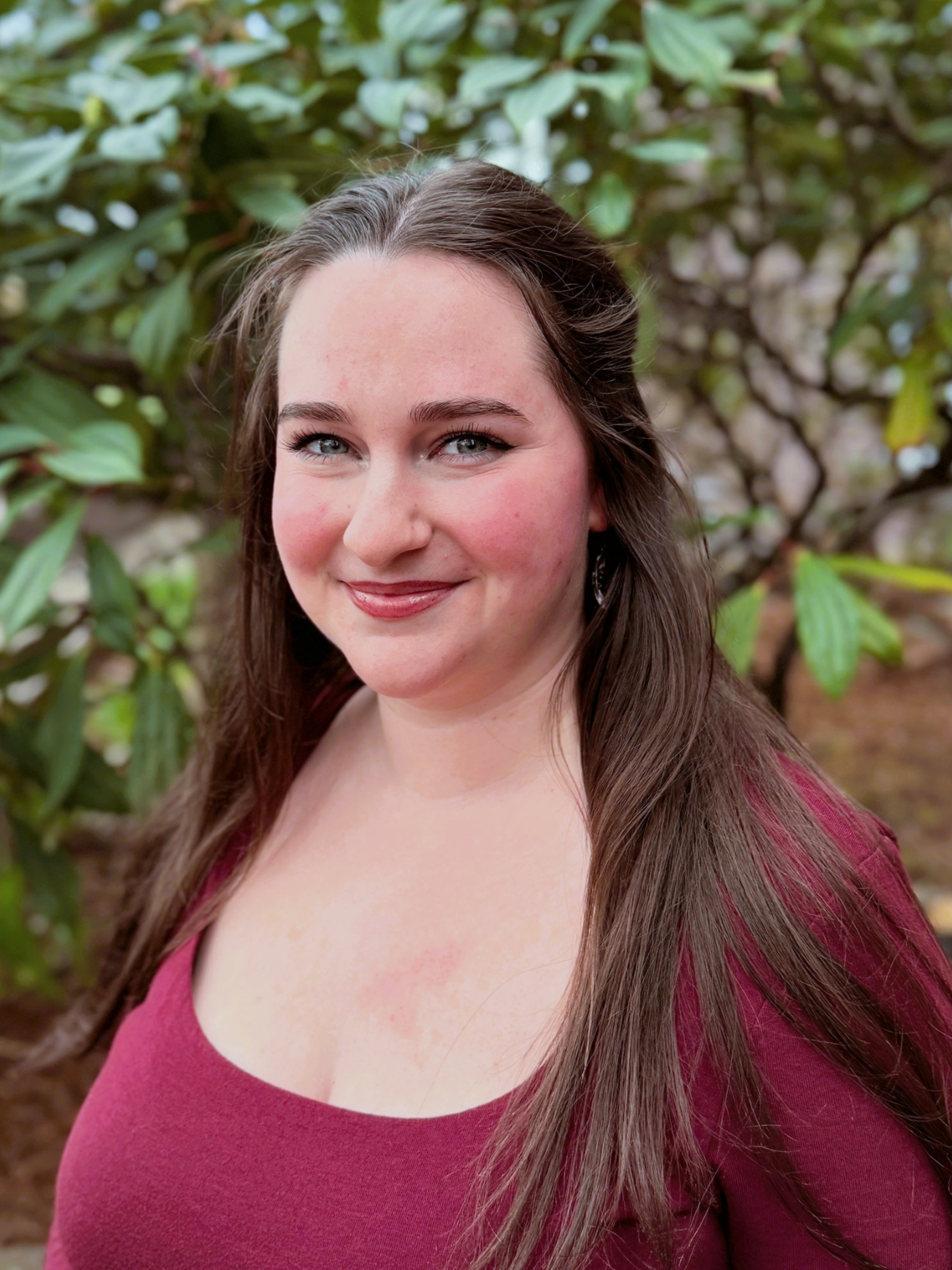 Close-up of a woman with long brown hair and blue eyes, smiling, wearing a burgundy top, outdoors with green leaves in the background.
