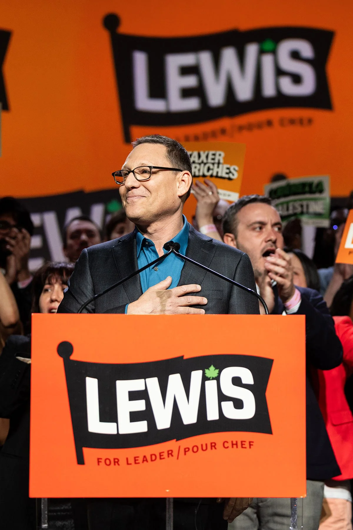 Avi Lewis speaks at a podium with the Lewis for Leader logo and a crowd of supporters behind him, against a logo step and repeat backdrop