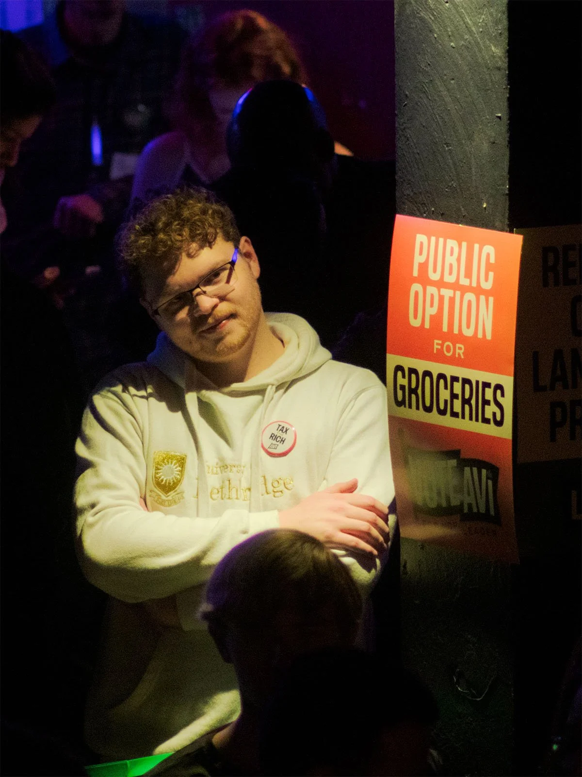 A supporter wearing a branded 'Tax the Rich' button leans agains a column with branded signage as they watch Avi Lewis speak at an event