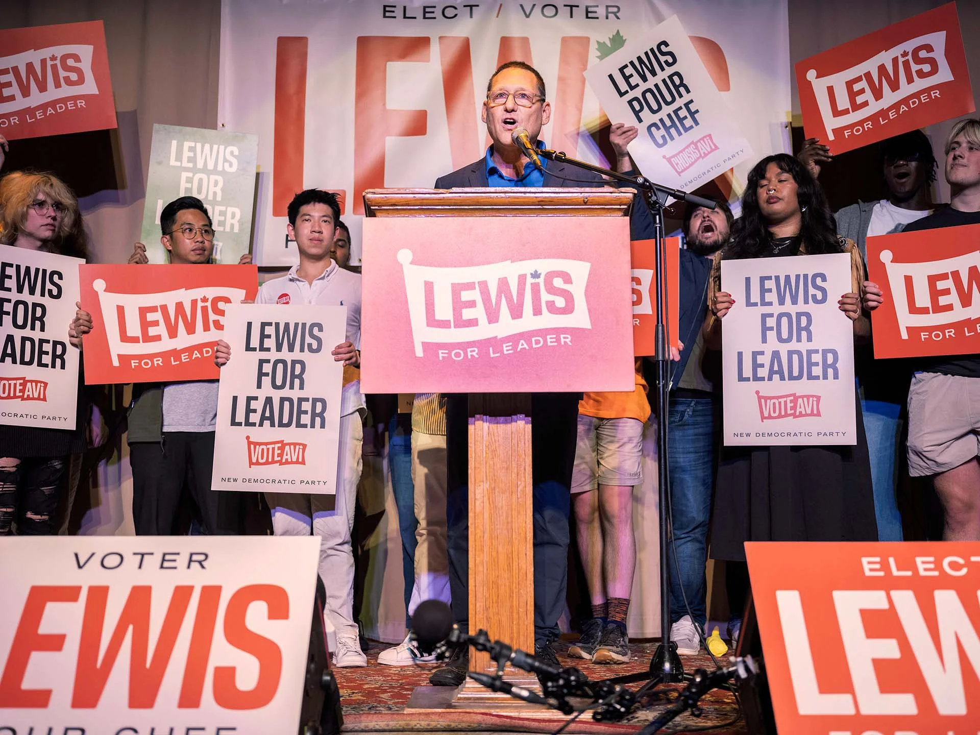 Avi Lewis speaks at a podium with the Lewis for Leader logo with supporters holding branded signage and a large branded vinyl banner behind him