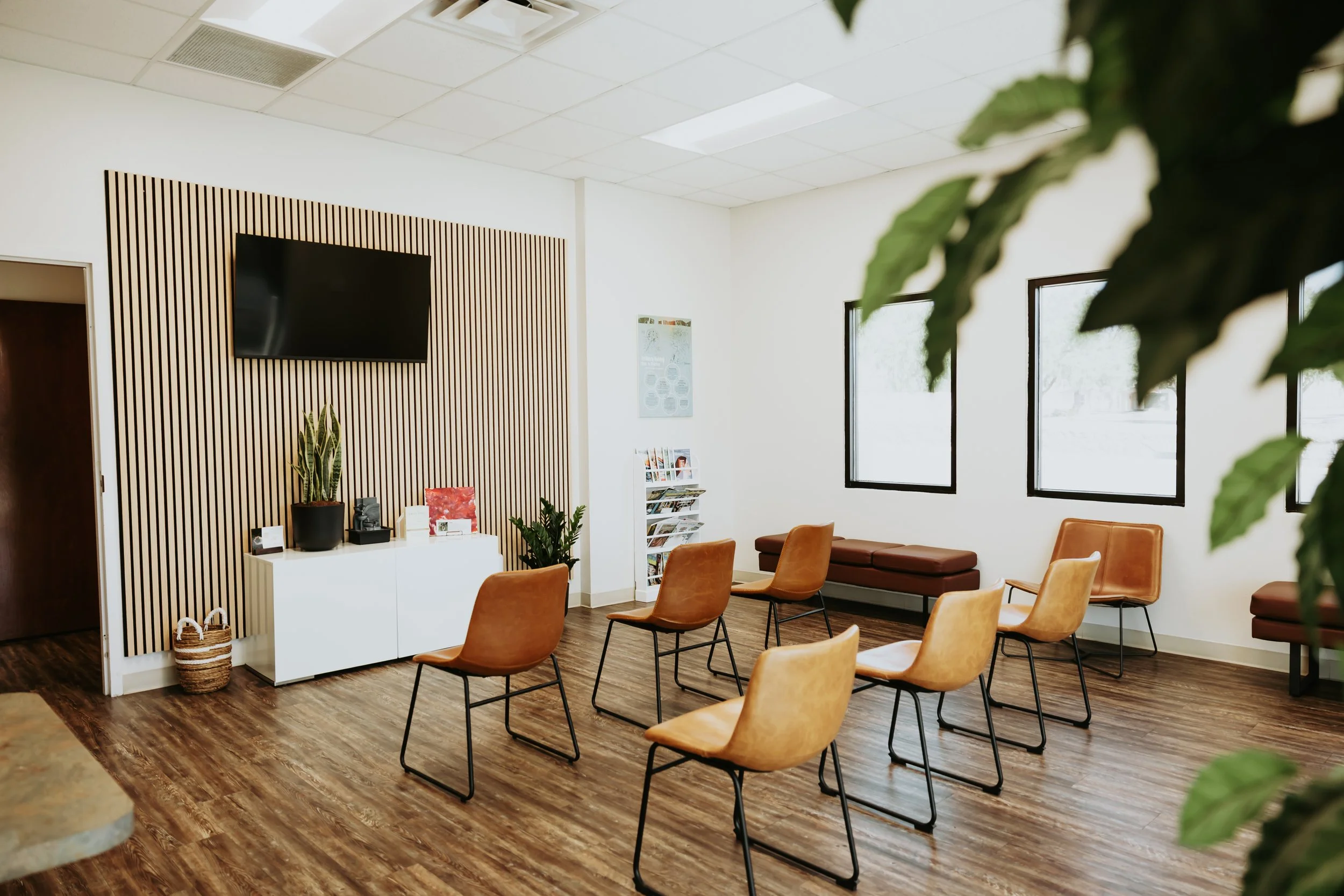 A waiting room with tan chairs arranged in rows, a white cabinet with plants and decorative items, and large windows letting in sunlight, with a slideshow on a wall and a magazine rack.