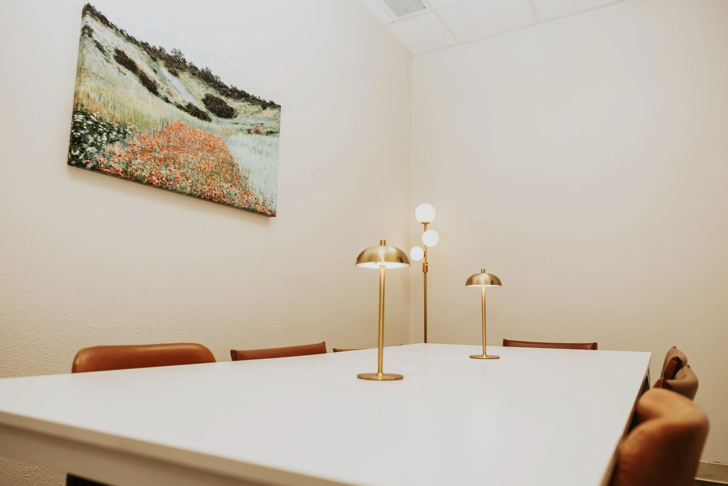A white conference table with brown chairs surrounding it in a minimalist room. Three gold and white table lamps are on the table. A landscape painting hangs on the wall and a modern lamp with multiple white globes stands behind the table.