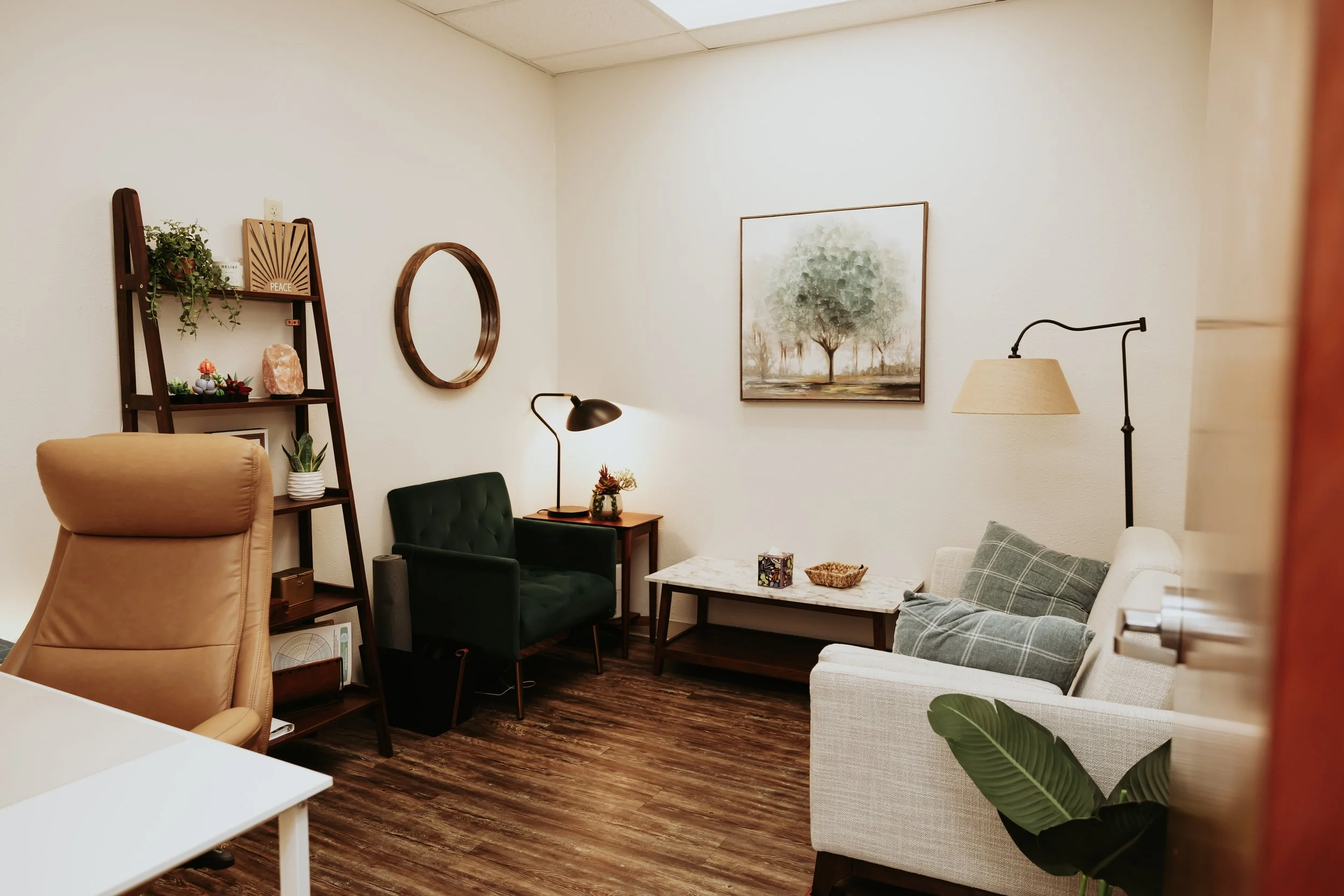 A cozy sitting room with a beige armchair, green velvet armchair, and a cream sofa with plaid pillows. Decor includes a bookshelf with plants and decorative items, a round wooden mirror, a framed tree painting, and a marble-top coffee table with a sm