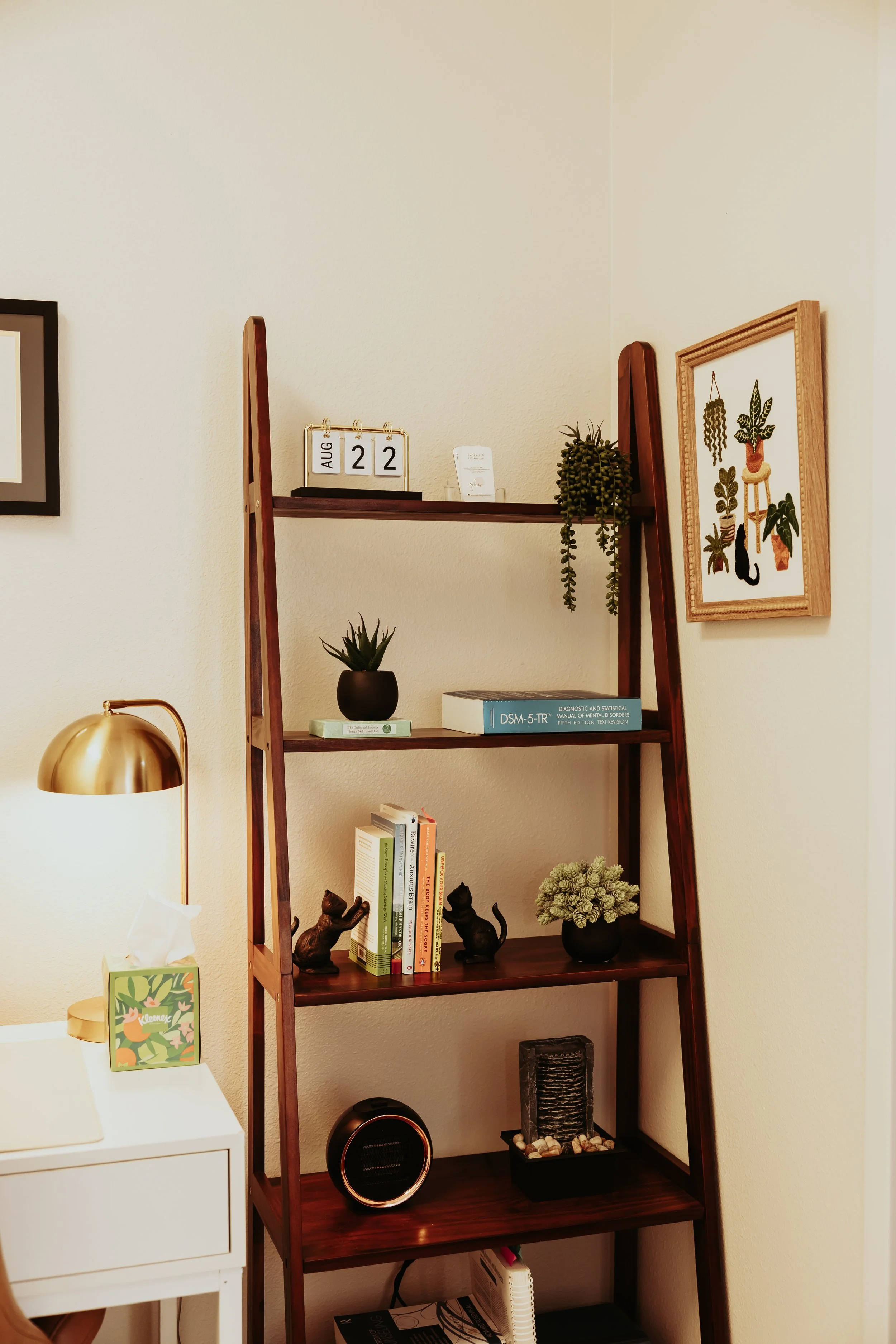 A wooden ladder-style bookshelf with four shelves in a living space. The top shelf has a small calendar showing August 22 and a business card holder. The second shelf has a potted succulent plant and a book about mental health. The third shelf has bo