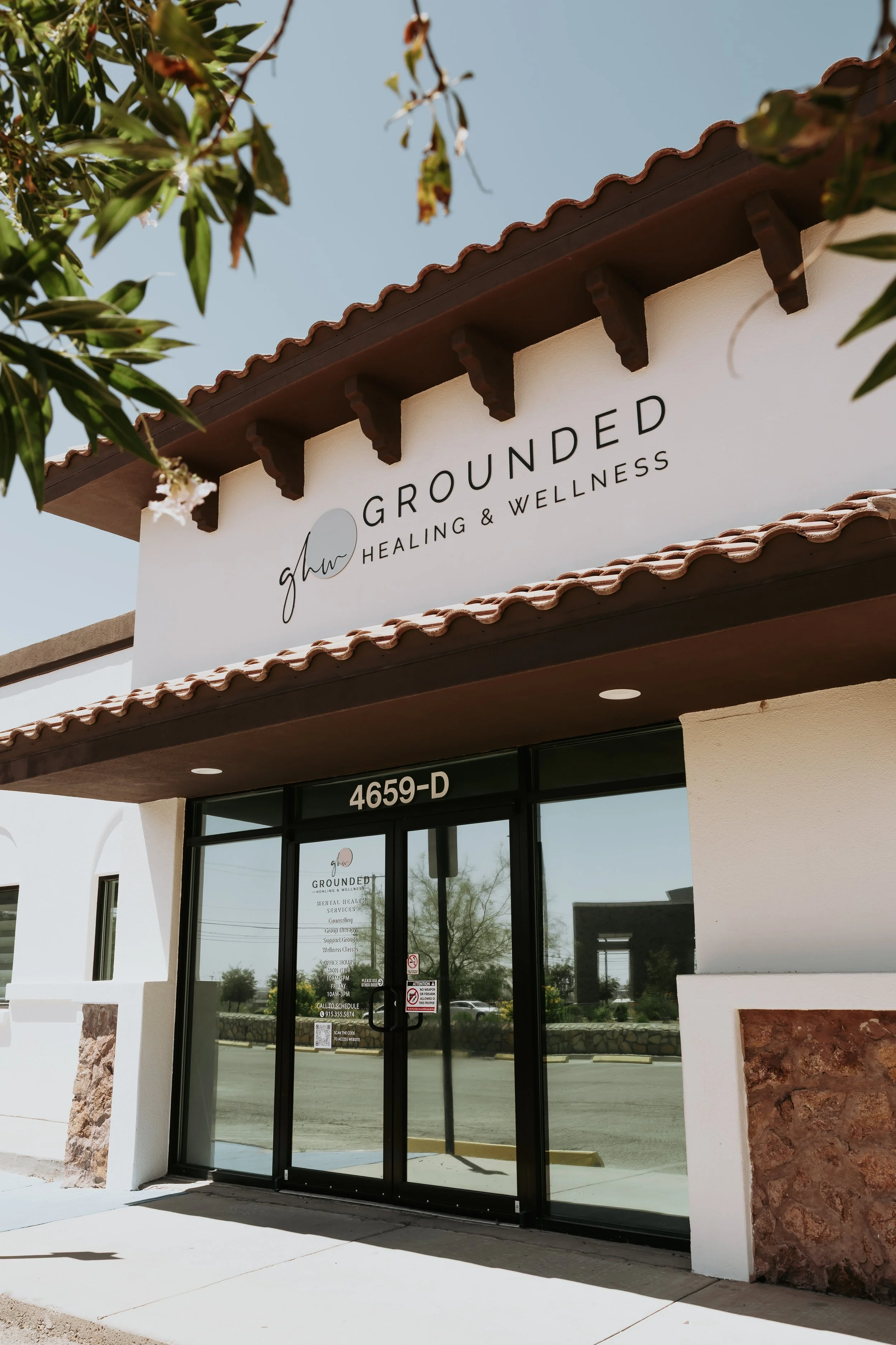 The storefront of Grounded Healing & Wellness, with a white sign featuring black lettering and a small logo, above glass double doors framed in black, with its street number 4659-D visible. The building has a tiled roof and a stone accent wall on the