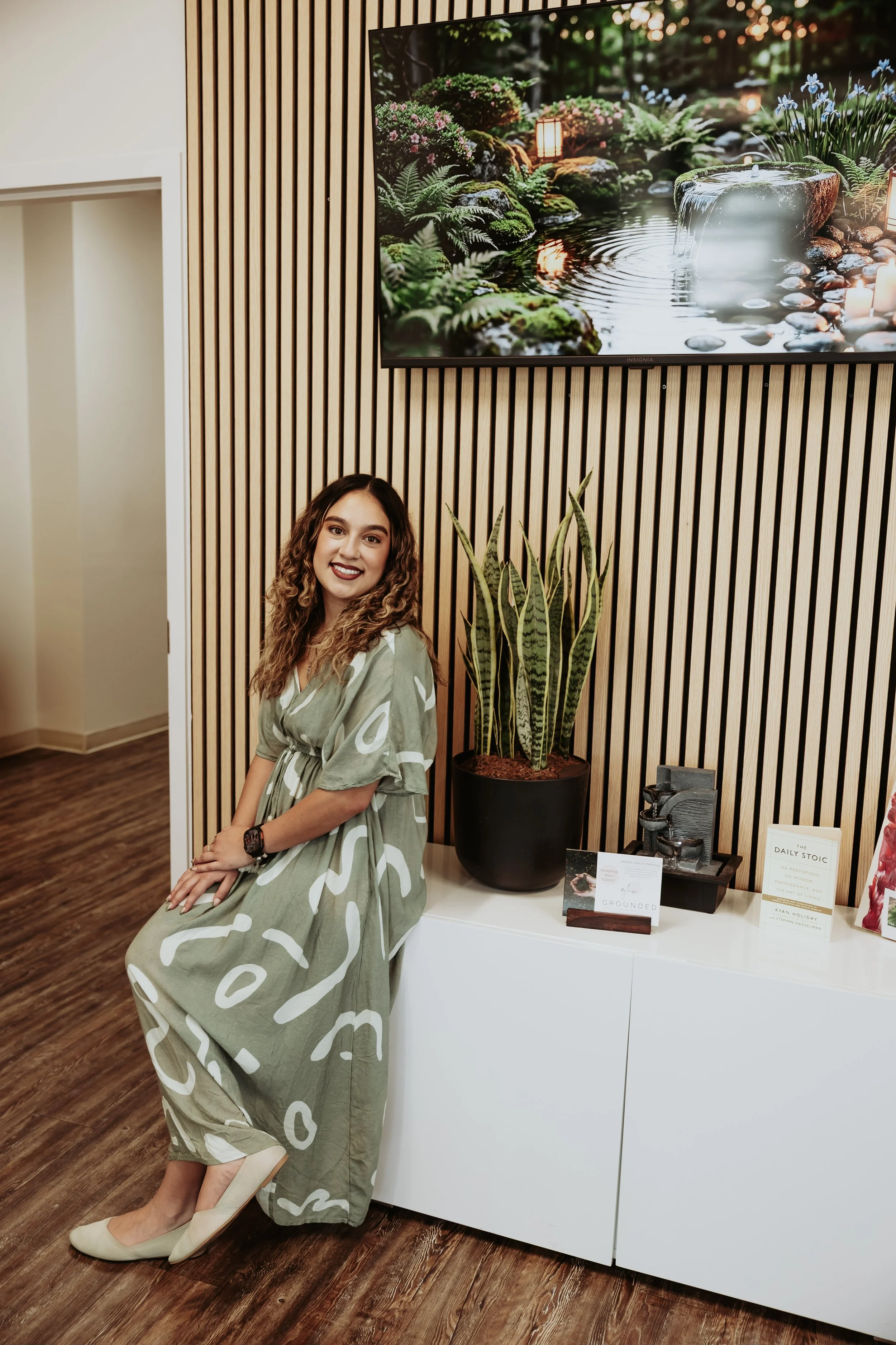 A young woman with wavy brown hair and a smiles sitting on a white stool in front of a large potted snake plant, with a wooden slatted wall, a table displaying books and decorative objects, and a large flat screen TV showing a serene nature scene with water and rocks