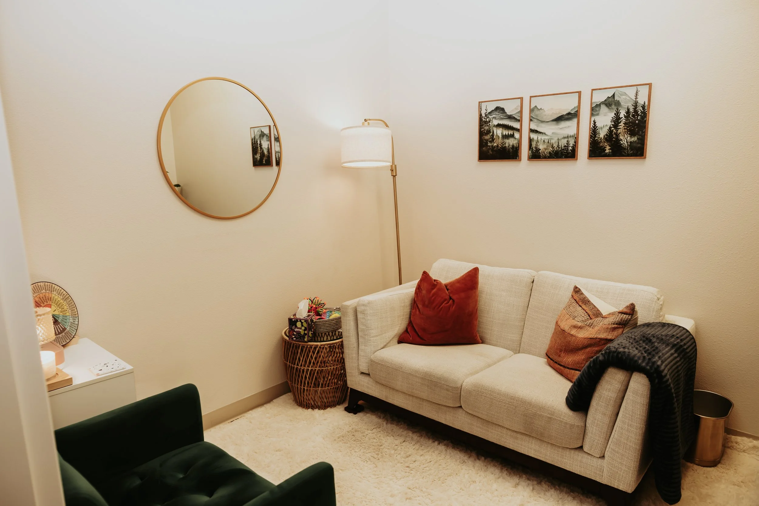 A cozy corner with a beige sofa decorated with brown and rust-colored pillows, a round wicker side table holding a colorful arrangement and tissue box, a tall floor lamp with a white shade, a large round mirror on the wall, and three landscape painti