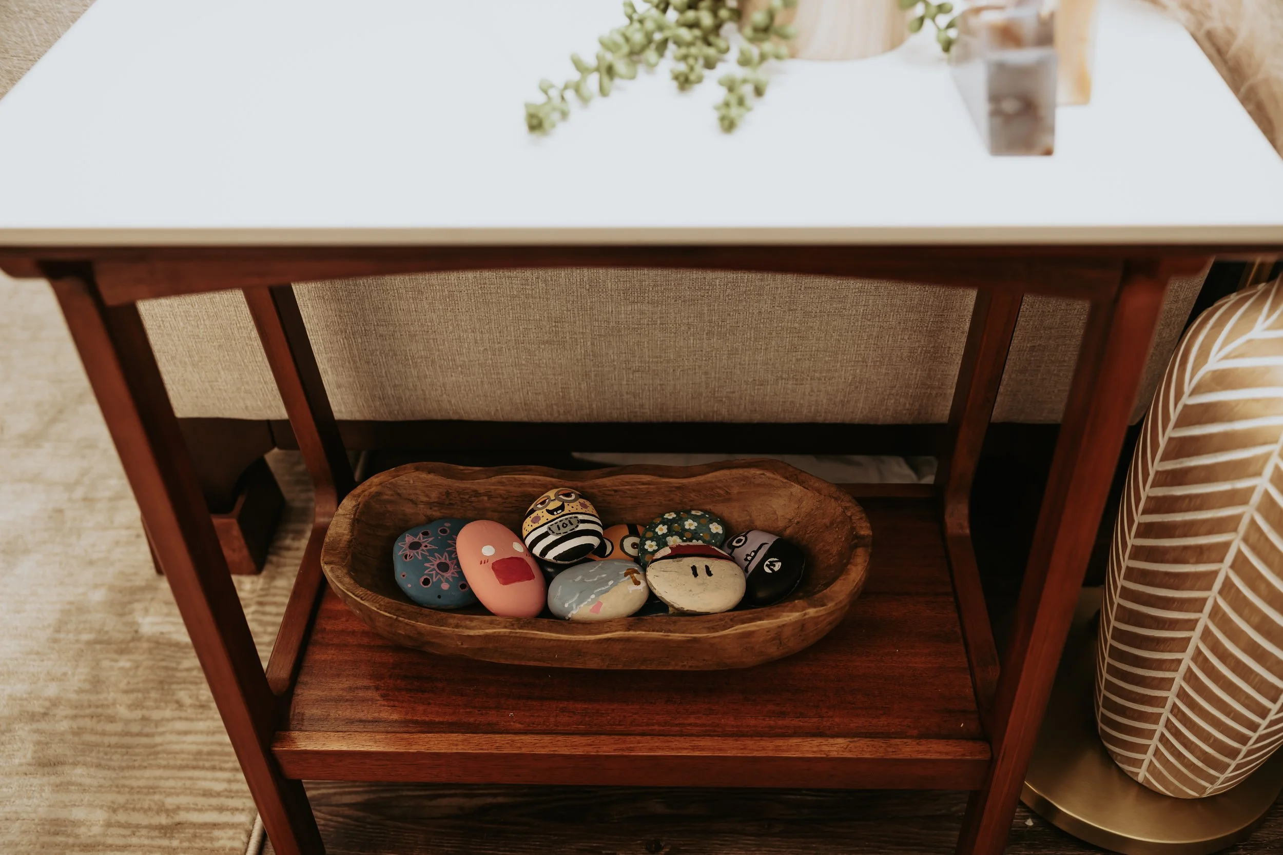 A wooden side table with a white top shelf and a lower wooden shelf holding painted rocks with designs, placed next to a striped armchair.