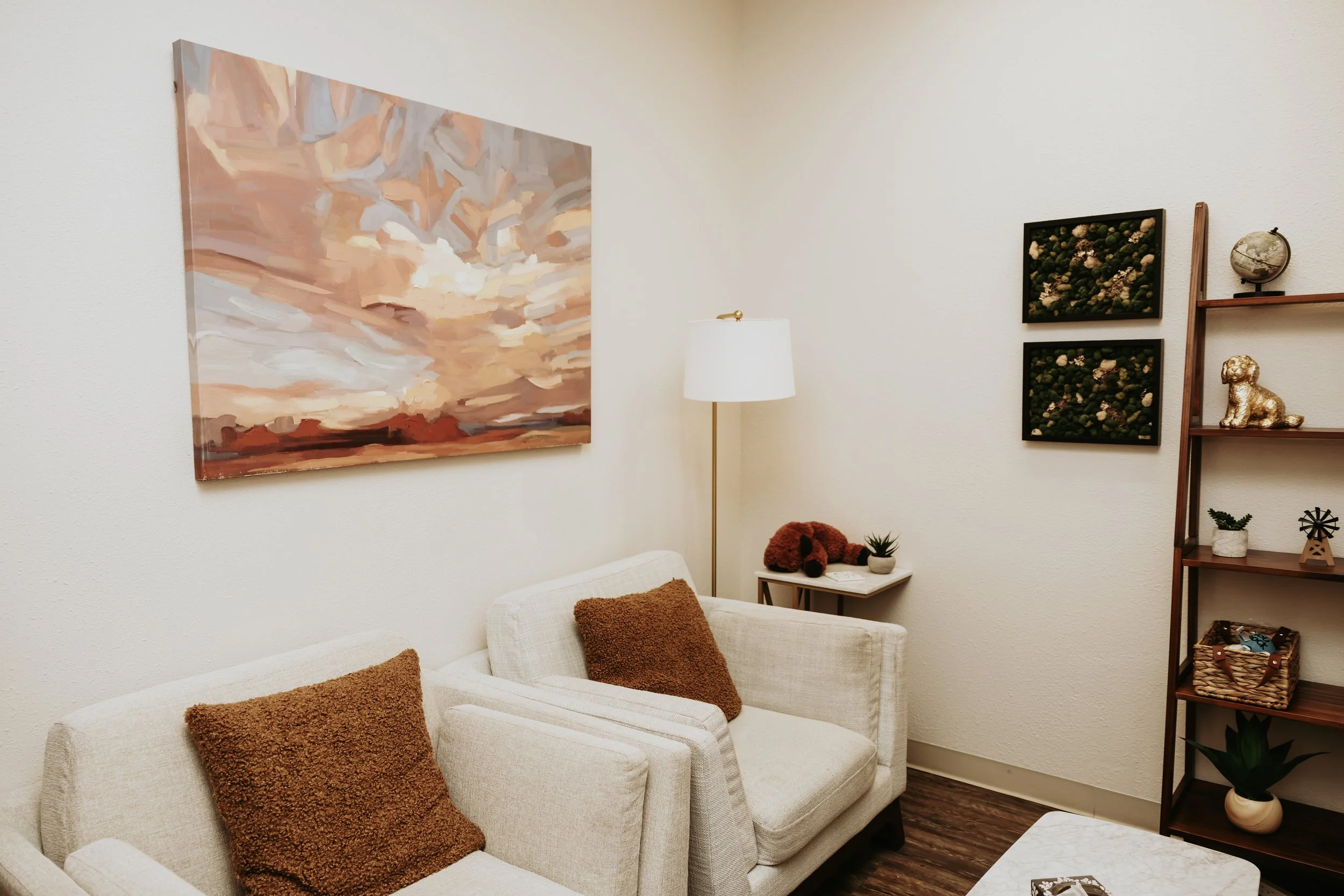 A cozy living room corner with a white sofa adorned with brown textured pillows, a tall white floor lamp, a wall gallery with framed art and decorative items including a globe and a small dog figurine, and a wooden bookshelf with plants, books, and d