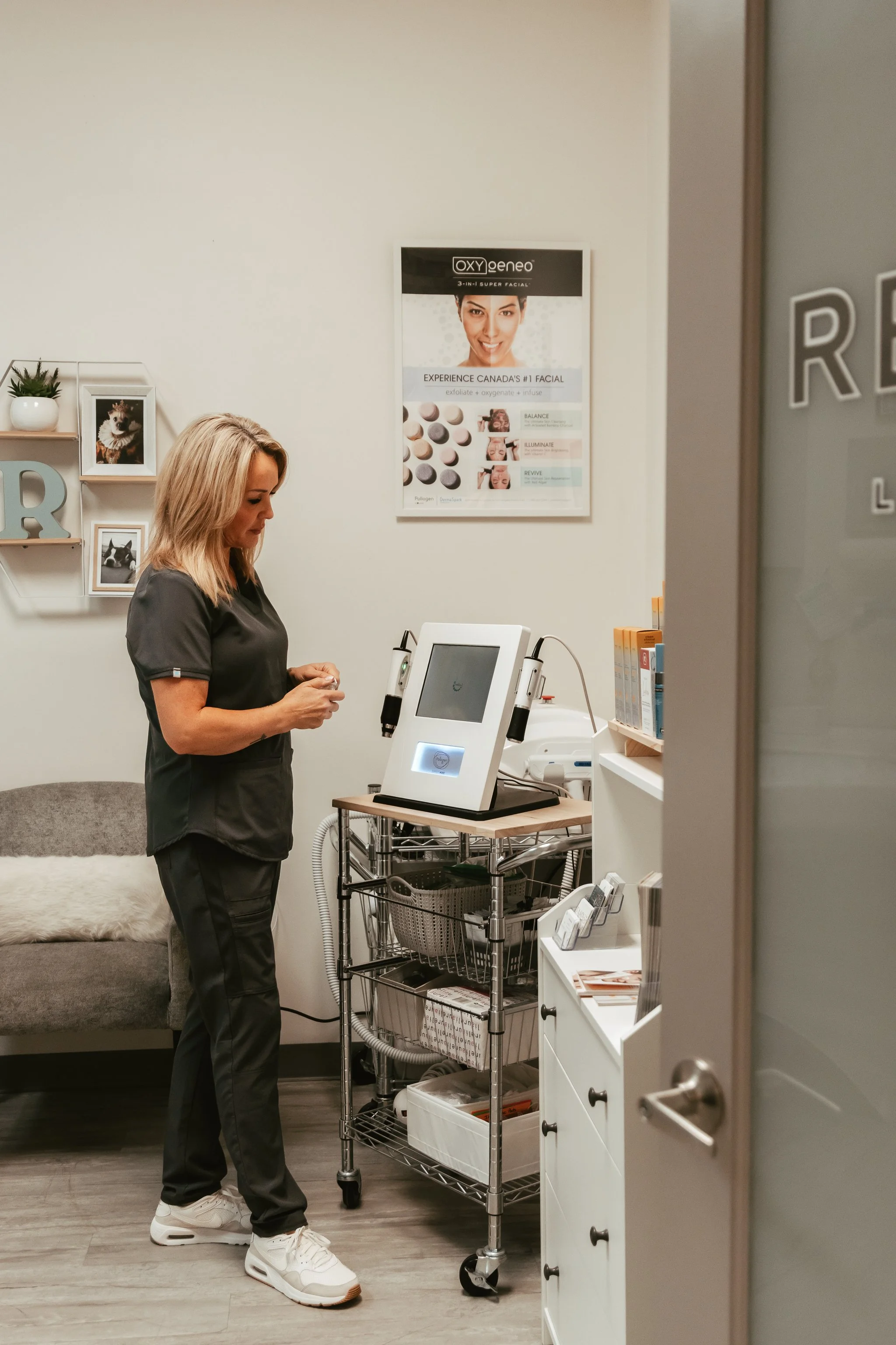 A woman in black scrubs looking at her phone in a medical office. Behind her is a gray couch with a blanket, a wall with framed pictures and decorative letters, and a medical device on a rolling cart. The door to the room is open, partially visible in the foreground.