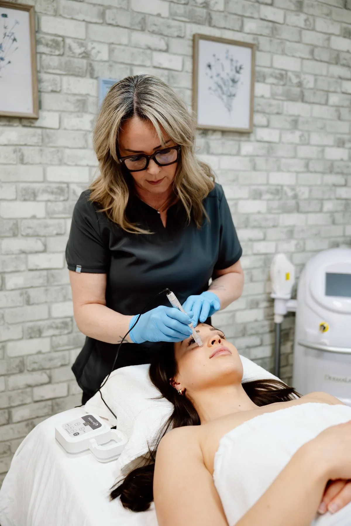 A woman receiving a cosmetic treatment on her eyebrows from a practitioner in a clinic with white brick walls.