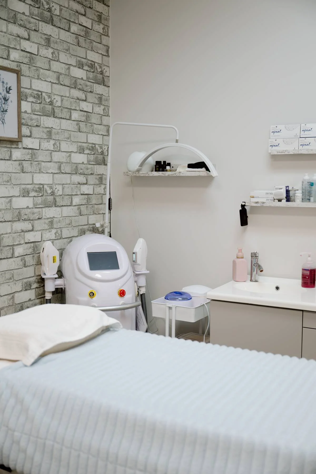 A medical treatment room with a white bed in the foreground, medical equipment including a machine with a screen and buttons, shelves with various medical supplies, a sink with soap and sanitizer bottles, and a brick accent wall on the left side.