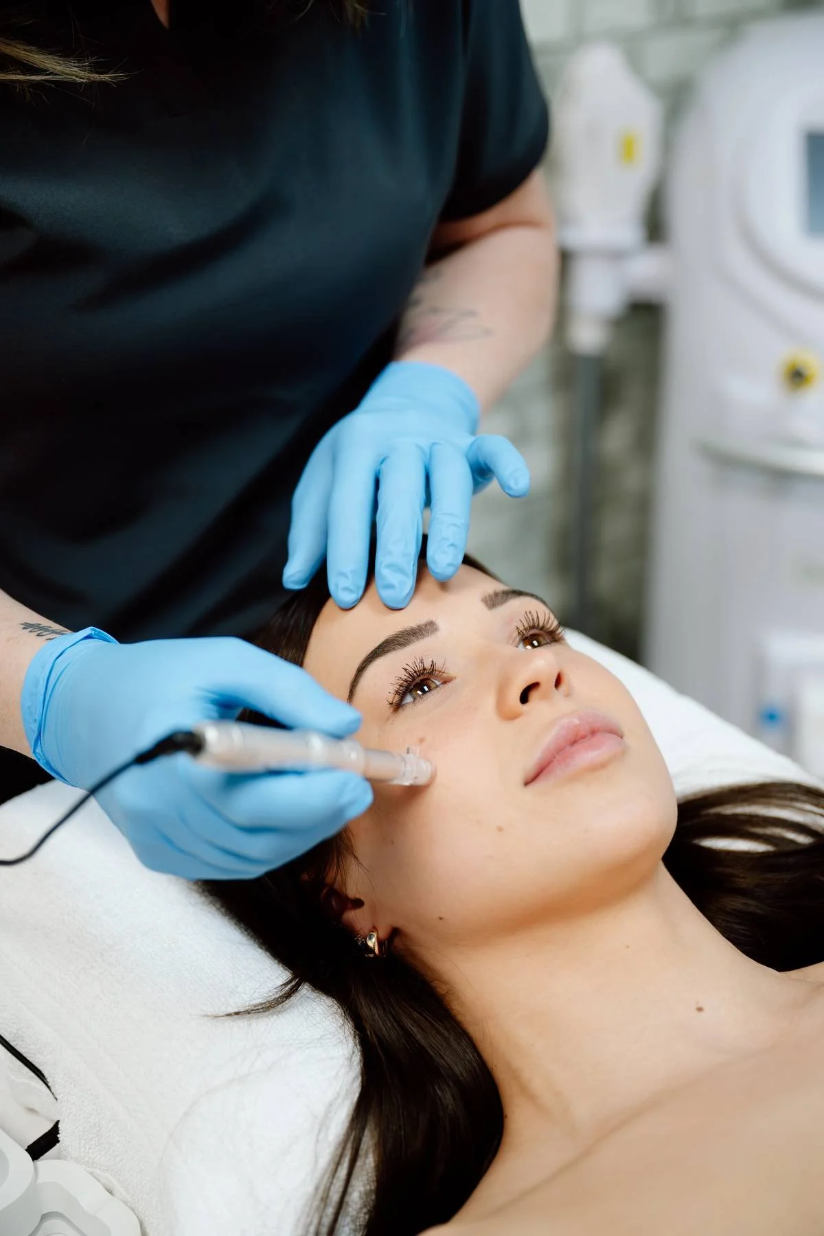A woman lying down on a treatment bed receiving a cosmetic facial injection from a healthcare professional wearing blue gloves in a clinical setting.