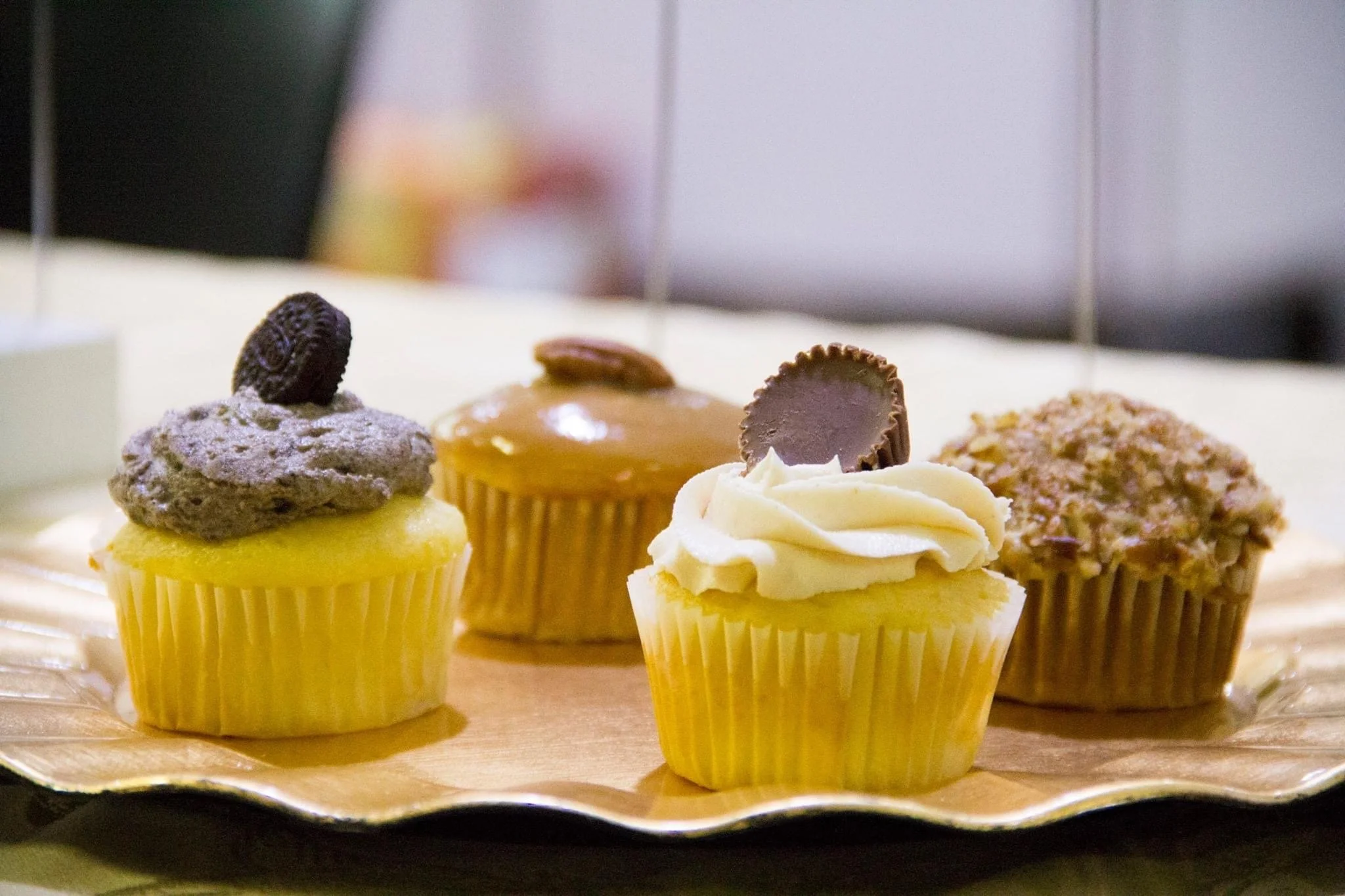 Four decorated cupcakes on a gold tray, featuring frosting, chocolates, and Oreo cookies.