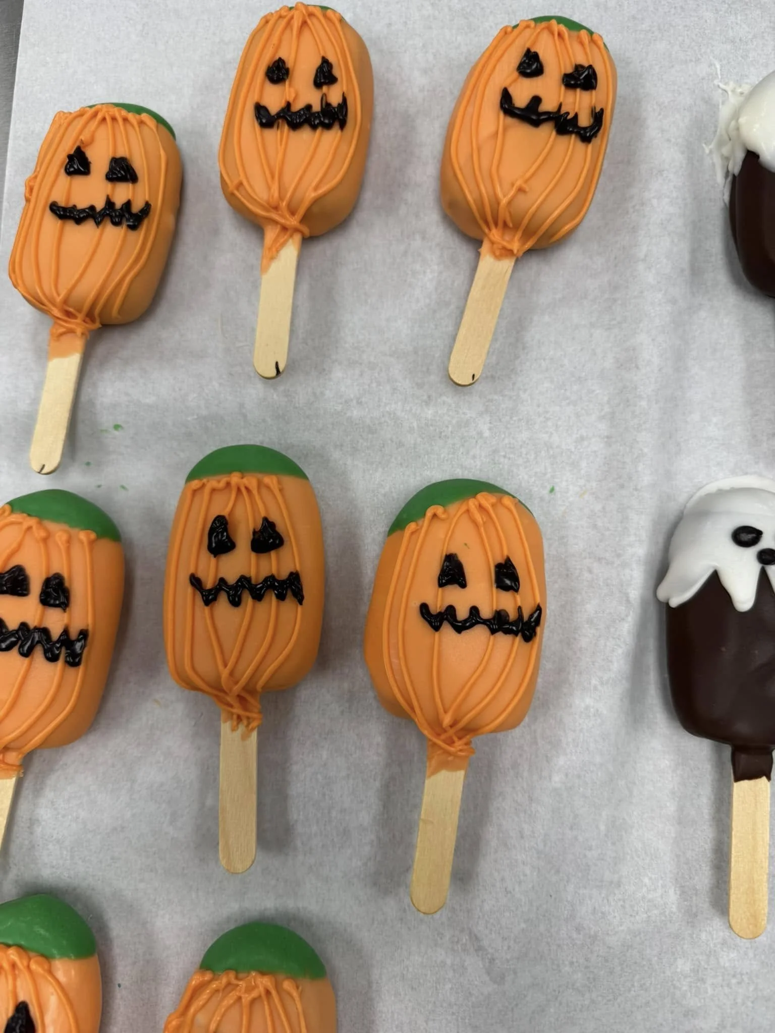Halloween-themed Cakesicles decorated as pumpkins with carved faces, green tops and orange drizzle