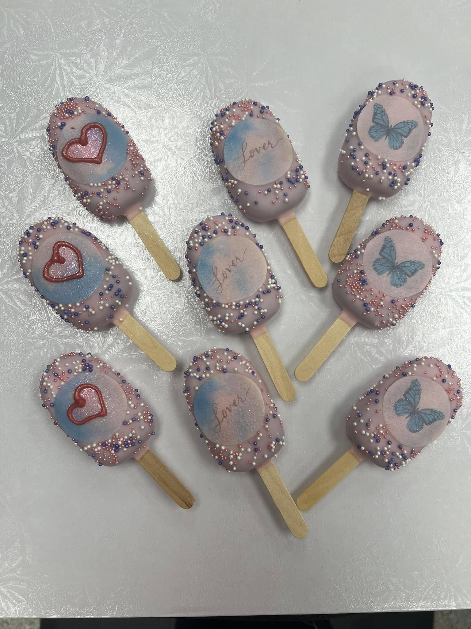 Pink Cakesicles decorated with multicolored sprinkles, some featuring blue butterflies and 'love' written on circular labels