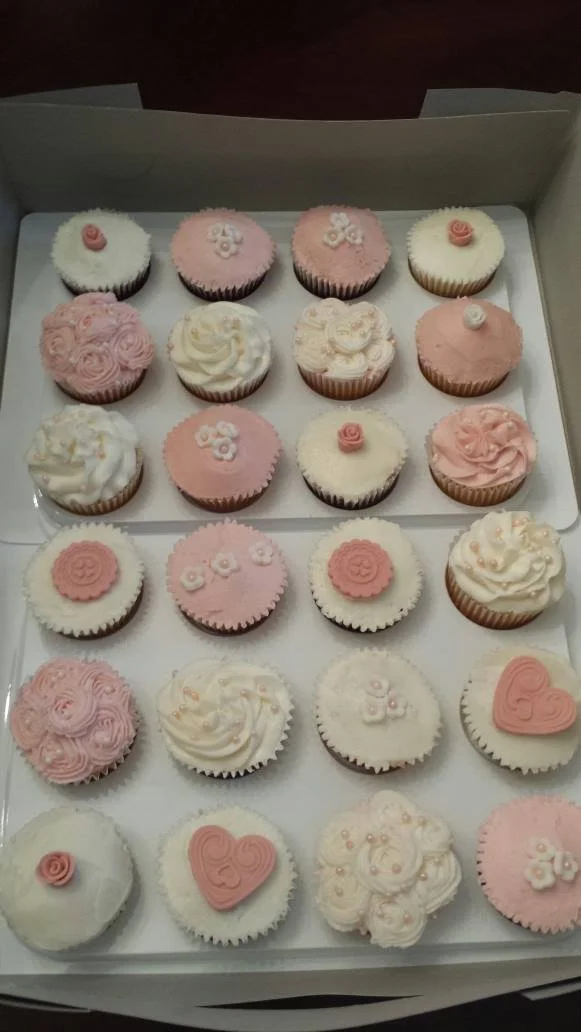 A box of assorted cupcakes with pink and white frosting, decorated with flowers, hearts, and swirls.