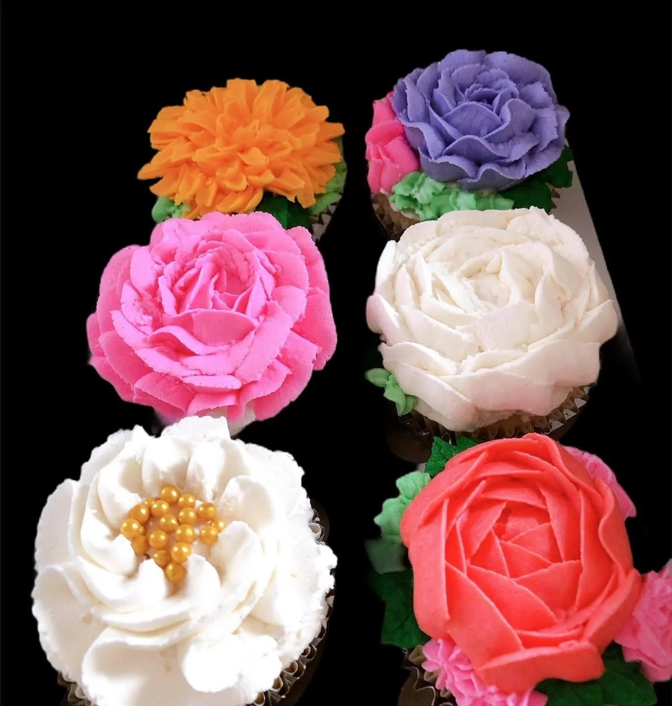 Six cupcakes decorated with colorful frosting and flowers, arranged on a black surface.