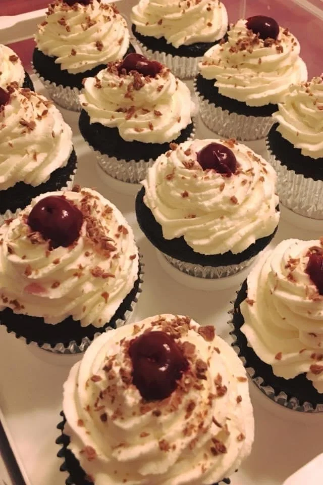 Cupcakes with white frosting, cherry on top, chocolate shavings, and crumbled cookie pieces in cupcake liners.