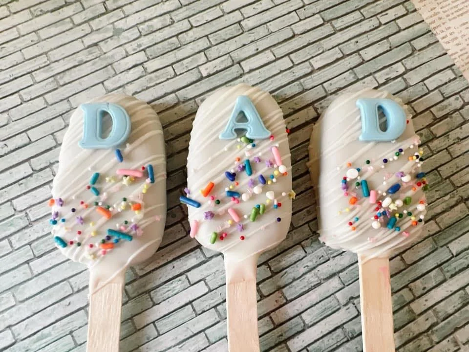 Three Cakesicles with white coating, decorated with multicolored sprinkles and blue fondant letter spelling 'DAD' 