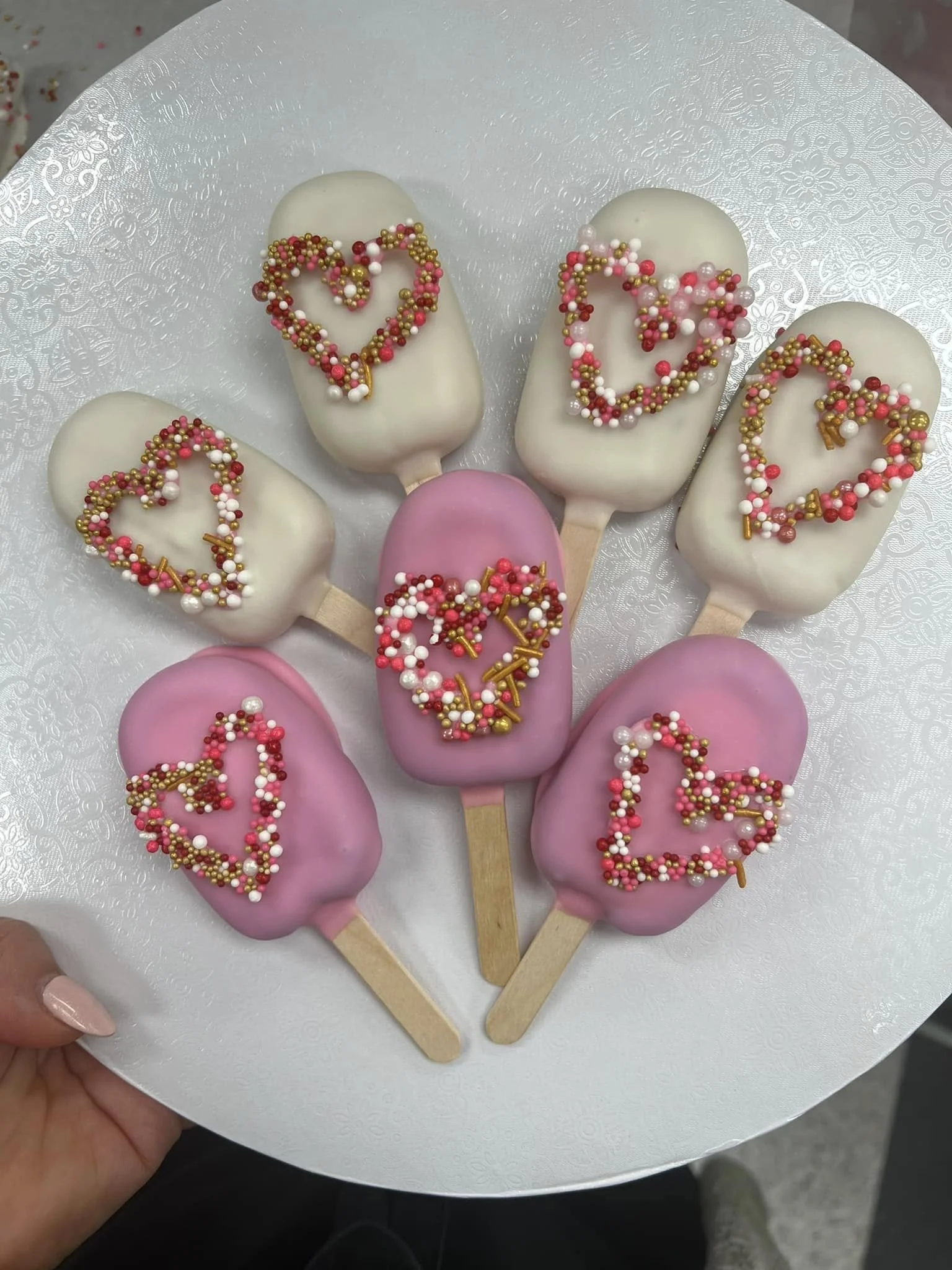 White and pink Cakesicles adorned with heart-shaped sprinkles in gold, pink, white, and red
