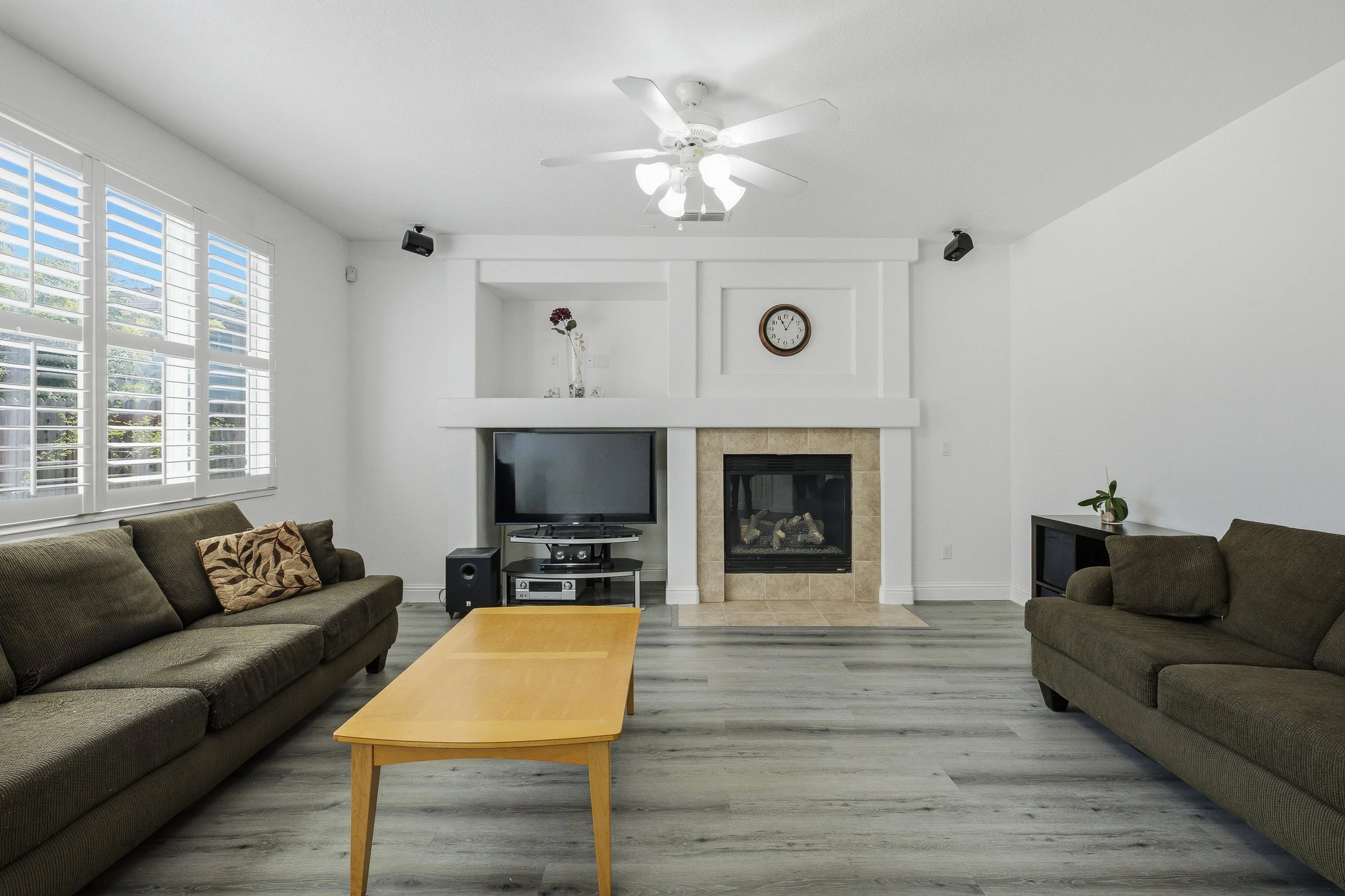 Living room with two brown sofas, a wooden coffee table, a fireplace, a wall clock, a TV, a stereo system, a vase with flowers, and window blinds.