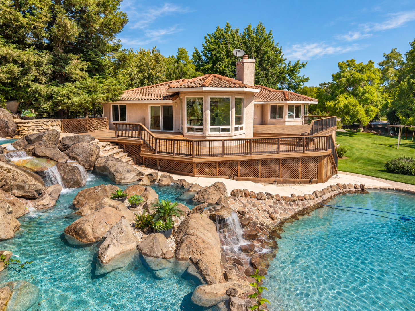 A house with a wooden deck overlooks a backyard with a large swimming pool surrounded by rocks, plants, and small waterfalls, with green trees and a blue sky in the background.