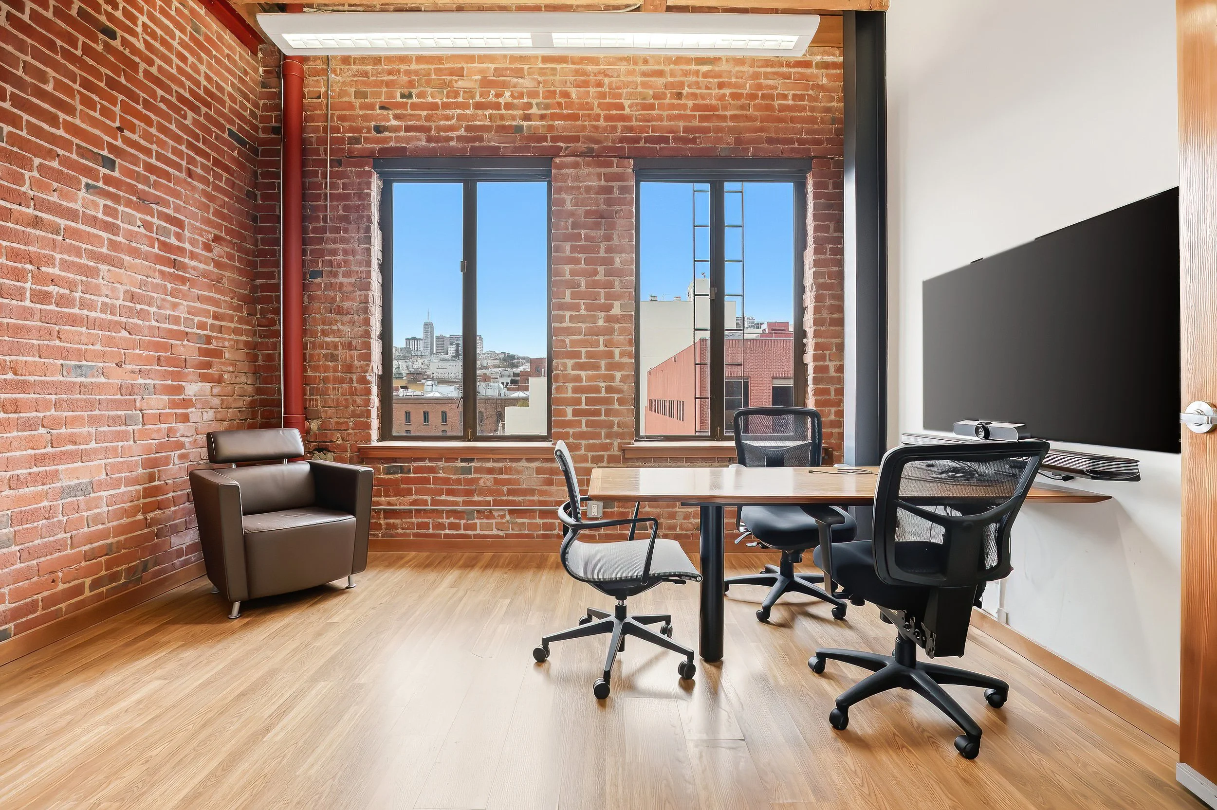 Modern office with exposed brick walls, large windows showing city skyline, wooden floor, a table with four office chairs, a large screen on the wall, and one armchair.