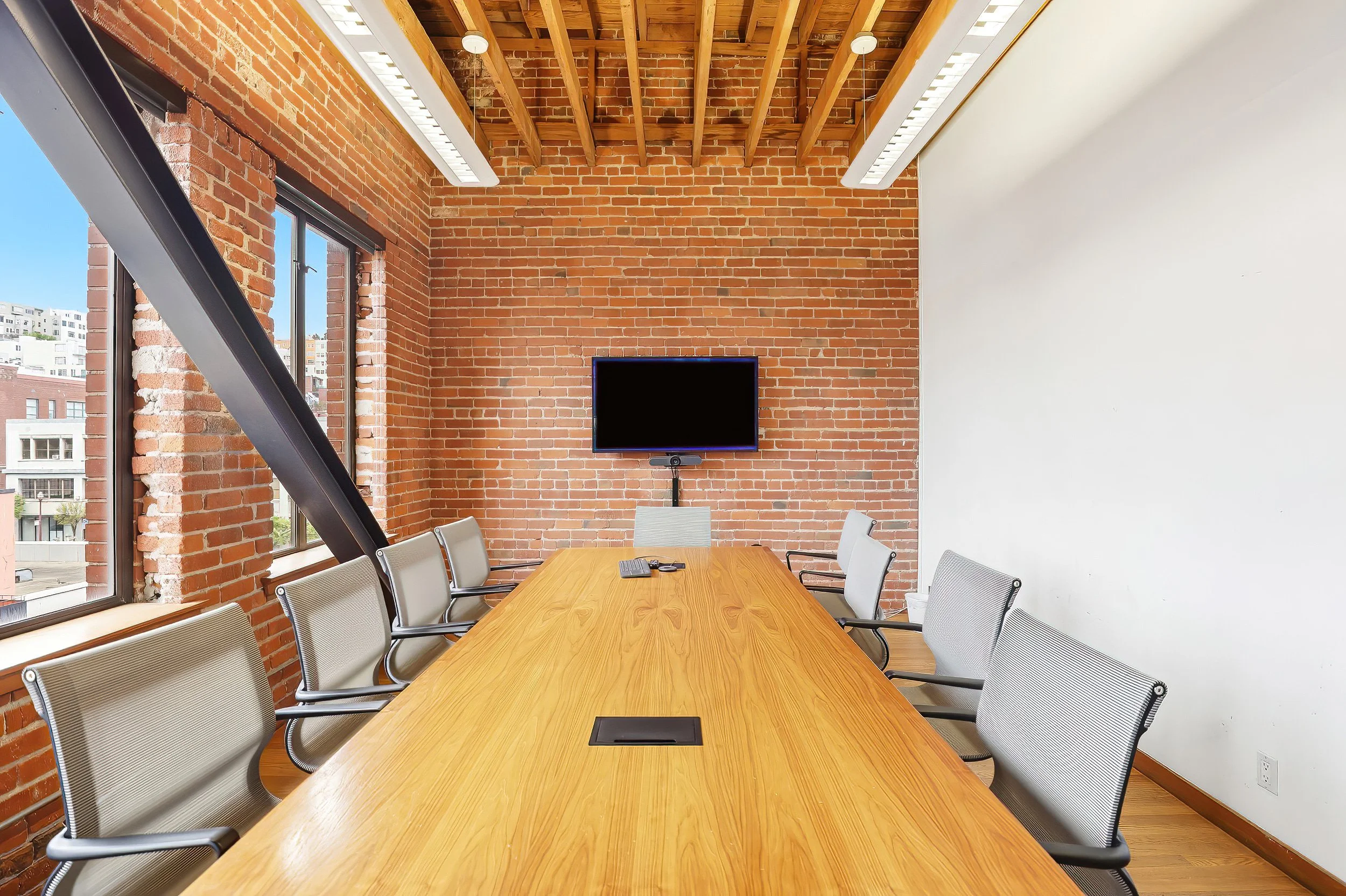 Empty conference room with a long wooden table, nine chairs, a wall-mounted TV, exposed brick walls, large windows, and a wooden ceiling with beams.