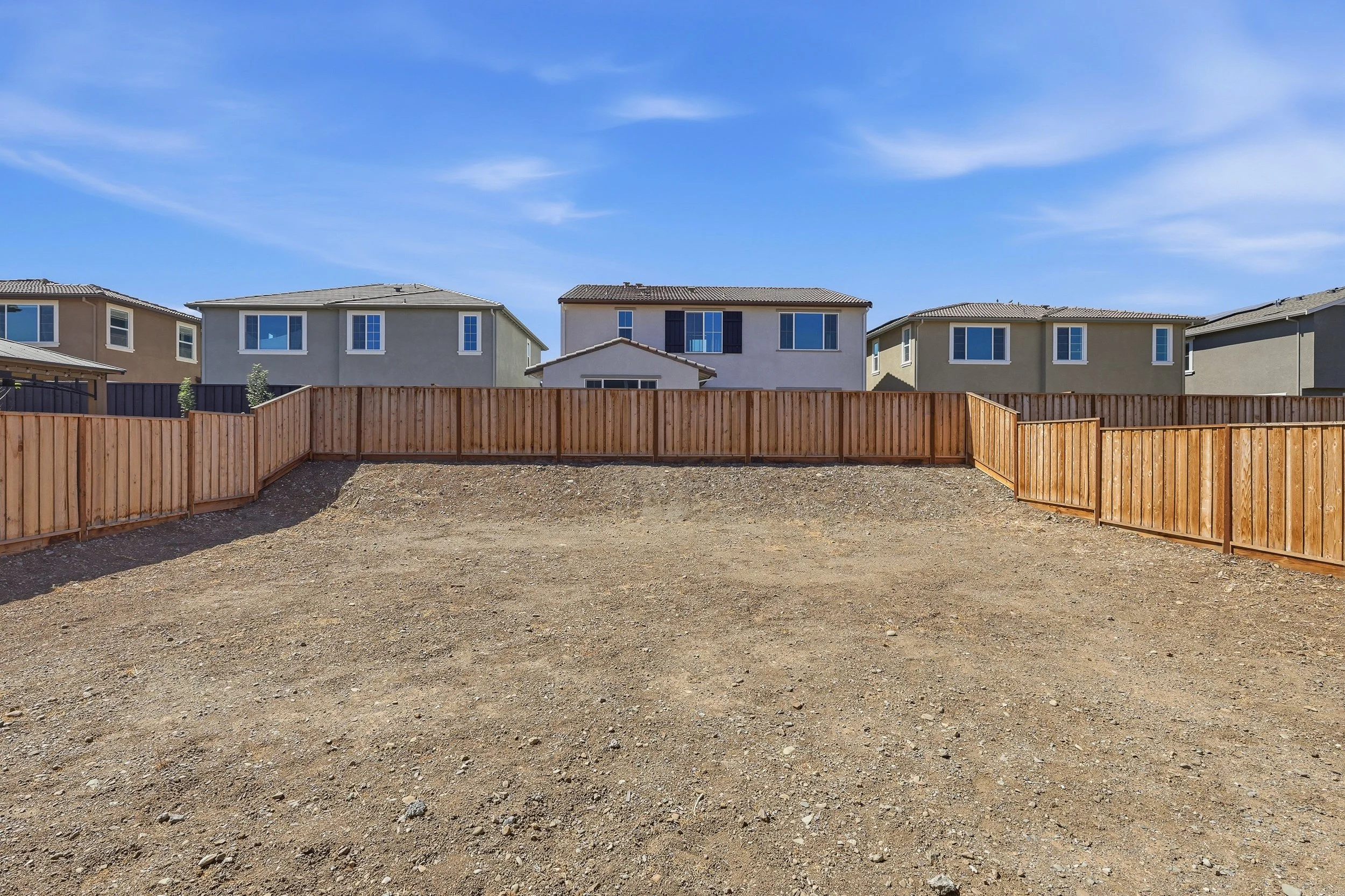 Fenced unfinished backyard with neighboring two-story homes beyond.