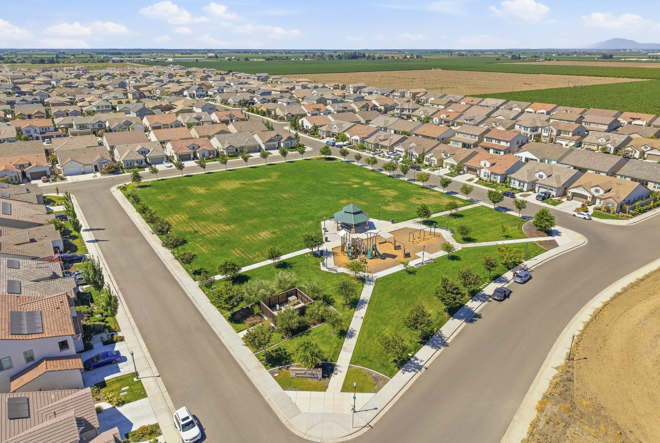 Aerial view of a suburban residential neighborhood with houses, a park, and surrounding farmland under a partly cloudy sky.