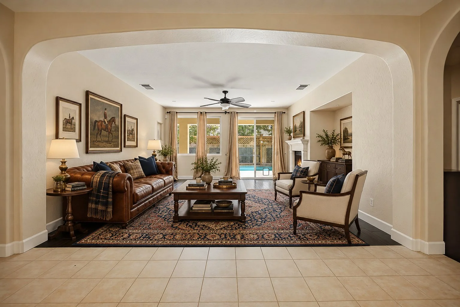 Living room with a leather sofa, two armchairs, a coffee table, a fireplace, hanging curtains, framed artwork, indoor plants, and a view of a swimming pool outside.