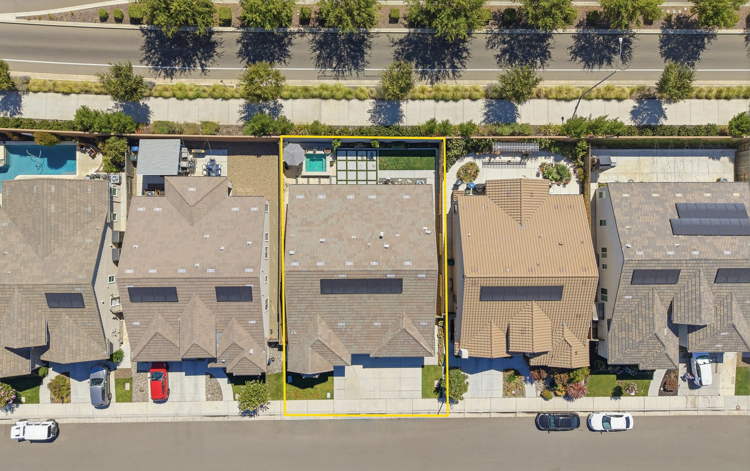 An aerial view of a residential neighborhood shows three houses with nearby yards, parked cars, a swimming pool, and a sidewalk lining the street. The middle house has a backyard with a patio, an umbrella, a small pool, and a green lawn, with trees a