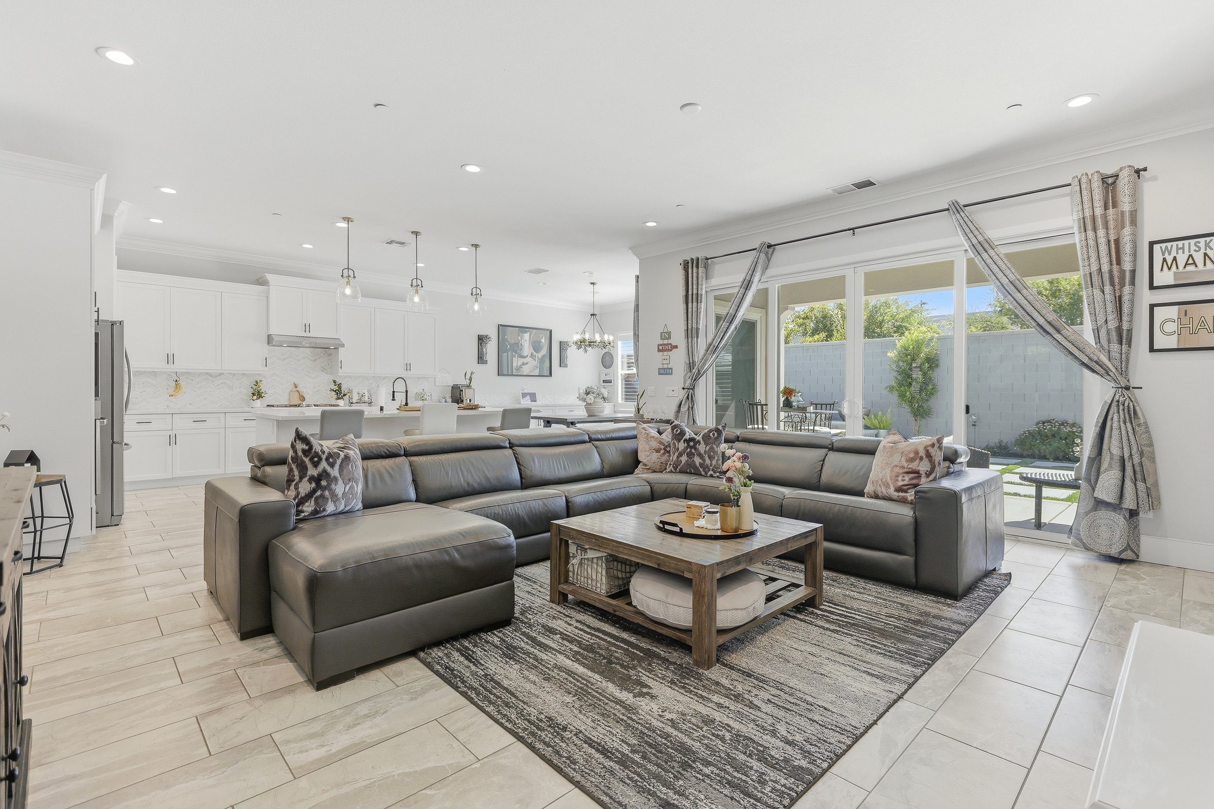 Open-concept living room and kitchen with large gray sectional sofa, wooden coffee table, patterned pillows, and bright sliding glass doors leading to backyard patio with outdoor seating and greenery.