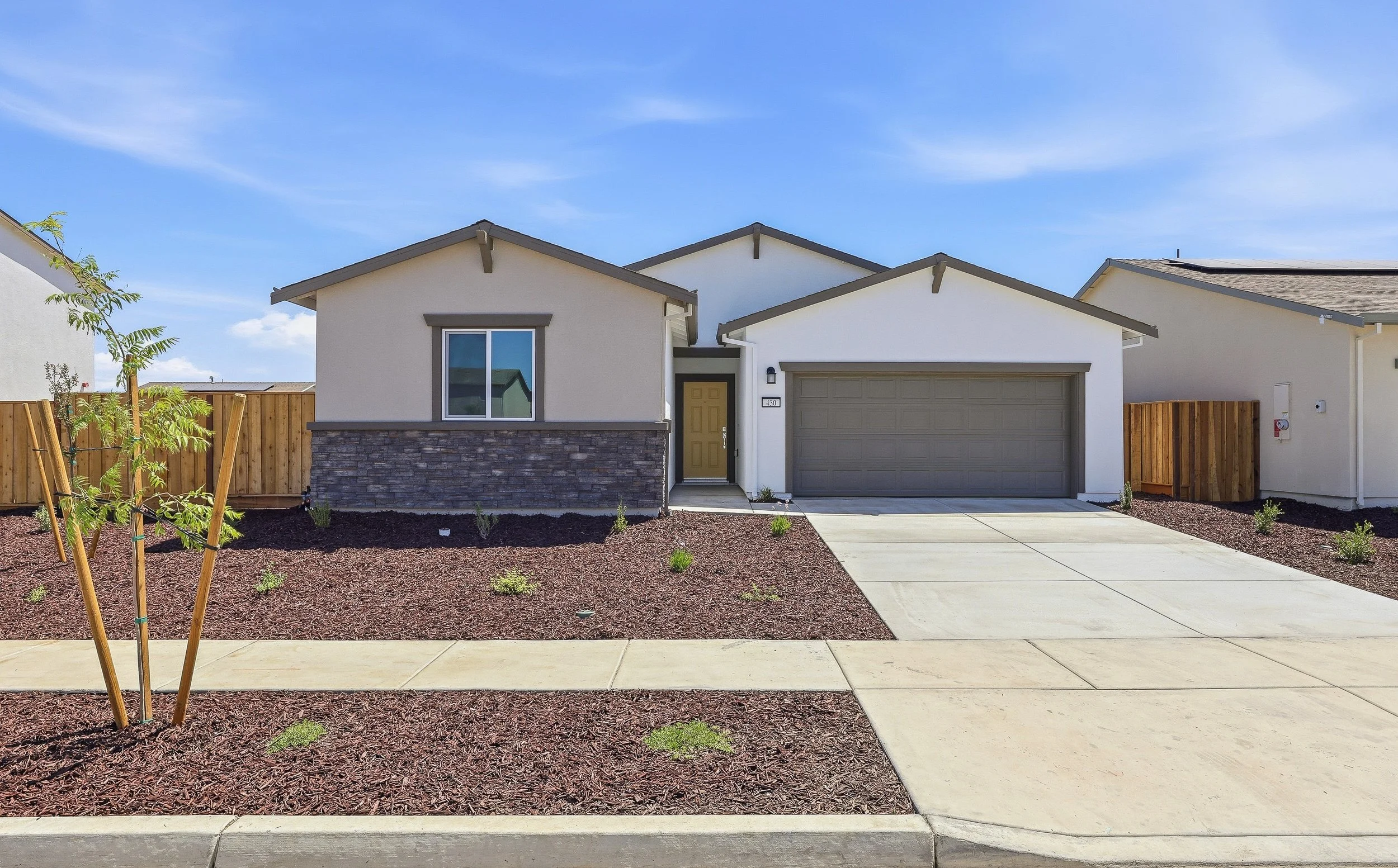 Modern residential house with a front yard featuring a young tree, mulch-covered garden beds, a concrete walkway, and a driveway. The house has white walls, stone accents, a yellow front door, and an attached garage. The sky is blue with some wispy c