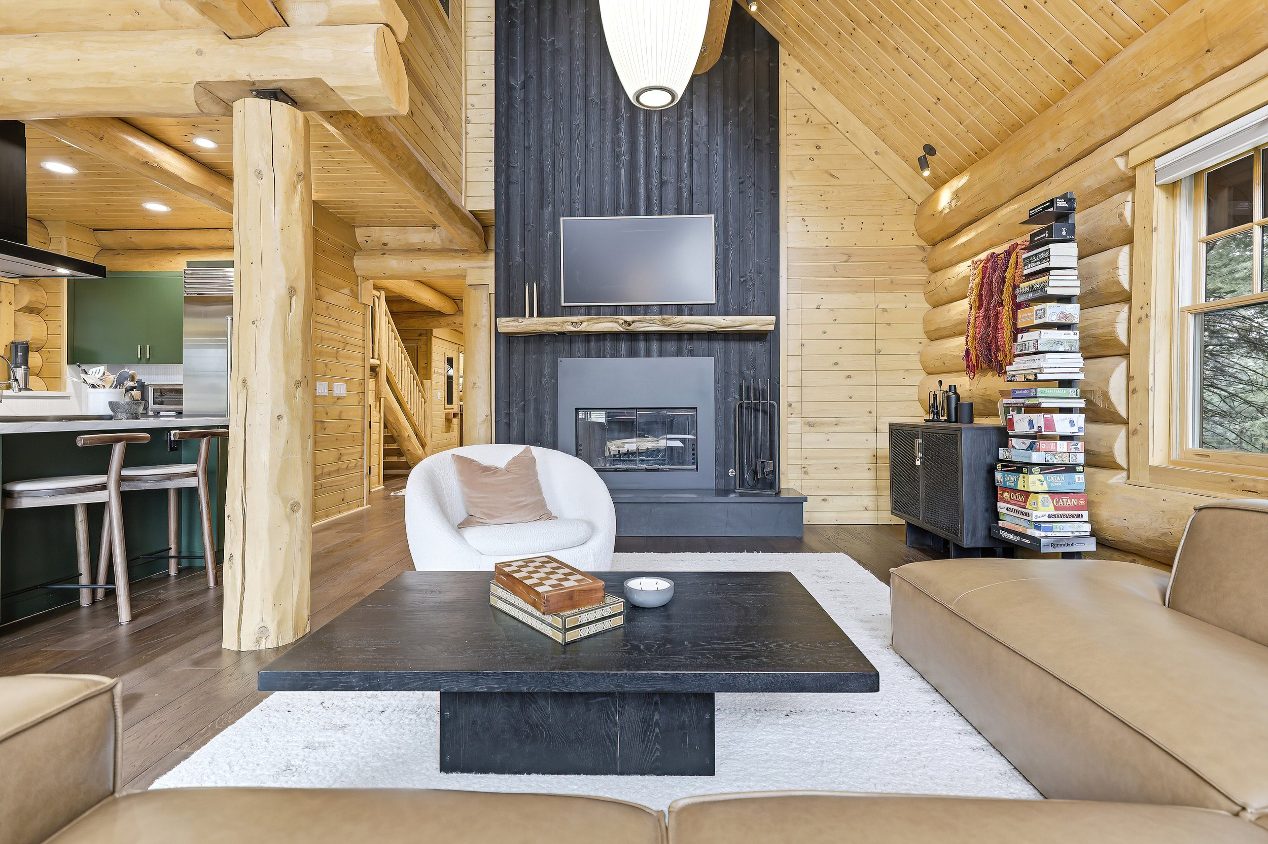 Living room with wooden walls and ceiling, fireplace, flat-screen TV, white armchair with a beige pillow, beige sofa, black coffee table with books and a candle, window showing trees, bookshelf with board games, ceiling light fixture, and a kitchen a