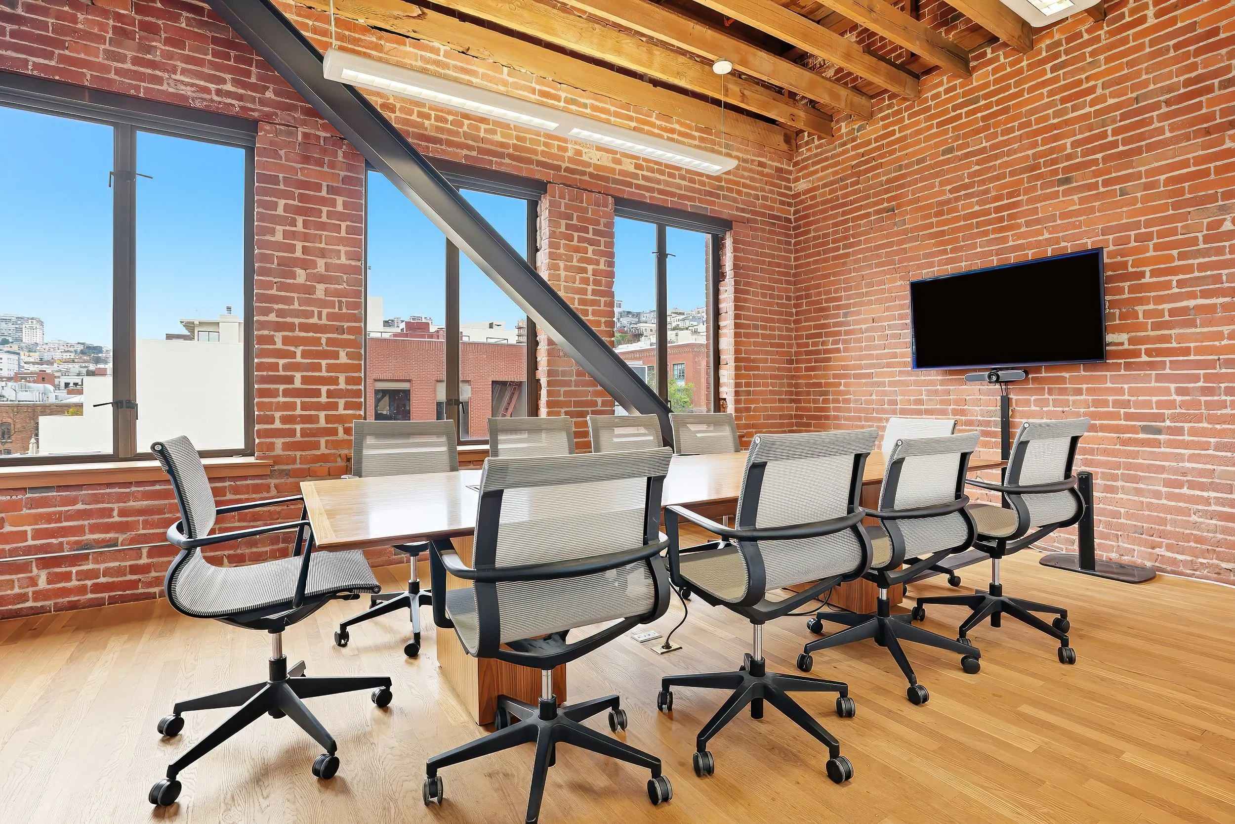 Conference room with brick walls, large windows, wooden floor, a long wooden table, nine grey office chairs, a flat-screen TV mounted on the wall, and ceiling beams with a light fixture.
