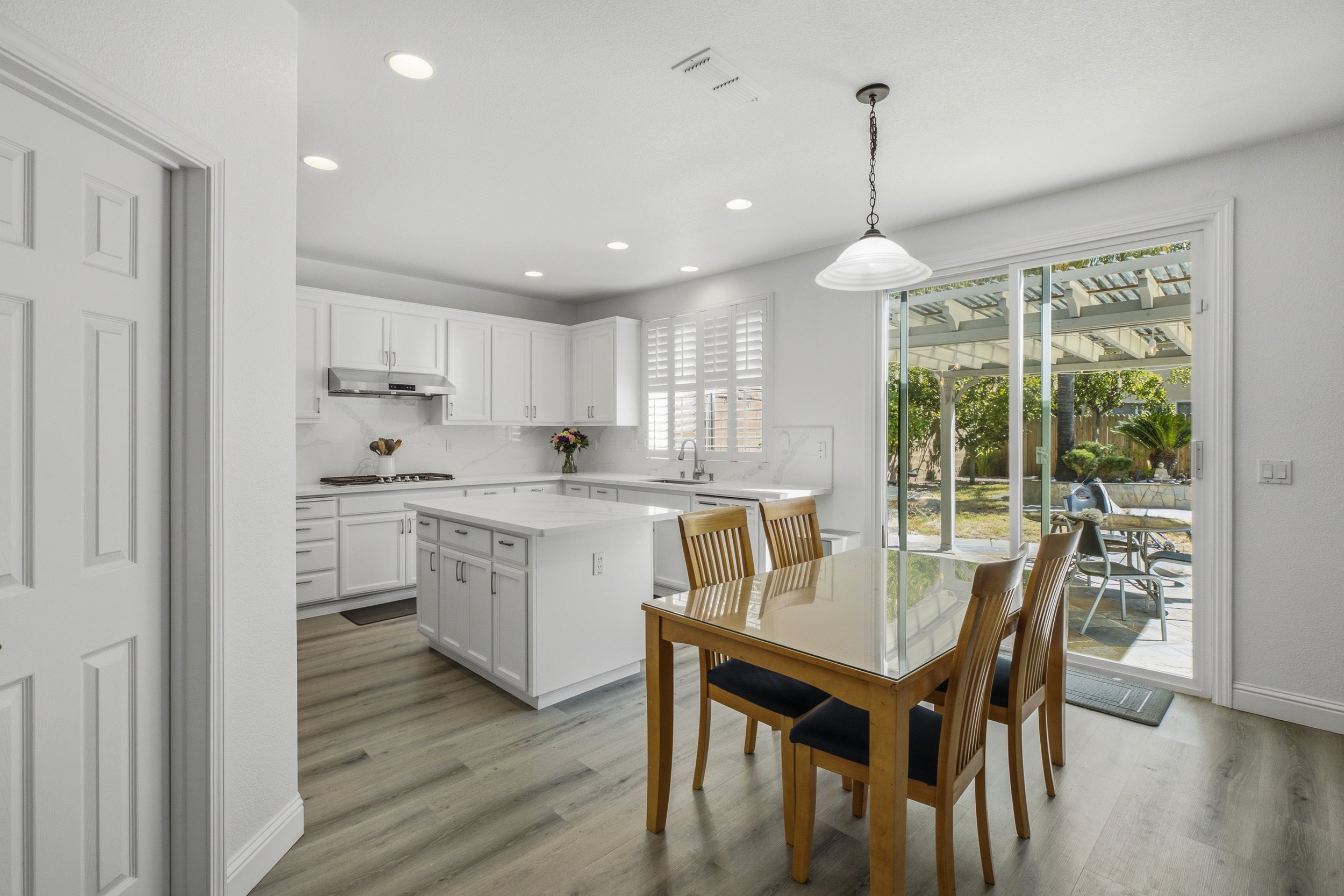 Kitchen with white cabinets, marble backsplash, large window with plantation shutters, sliding glass door leading to backyard, wooden dining table with four chairs, and a pendant light fixture.