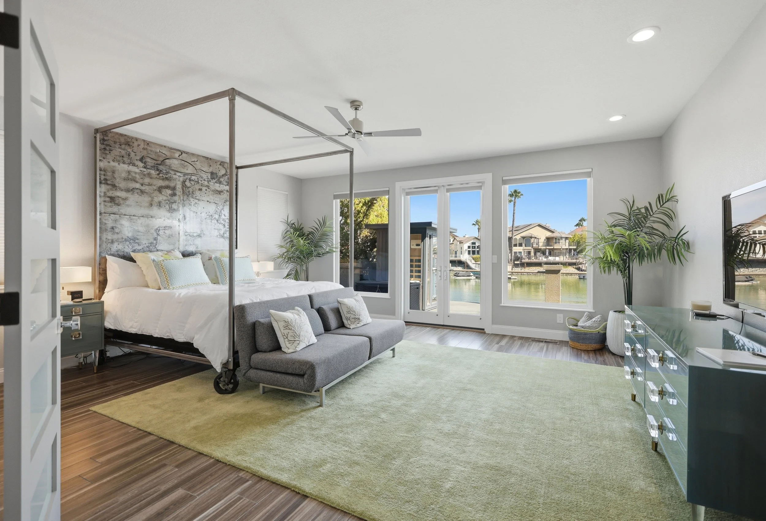 Living room with a bed, gray sofa, large windows showing a canal and houses outside, green plants, and furniture including a TV and a green rug.
