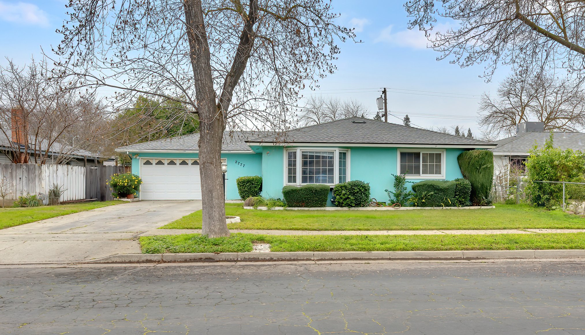 A light blue single-story house with a gray shingle roof, large front window, and a garage door. The house has a front yard with a tree, green bushes, and a small garden. A concrete driveway and sidewalk are visible in front, with a street in the for