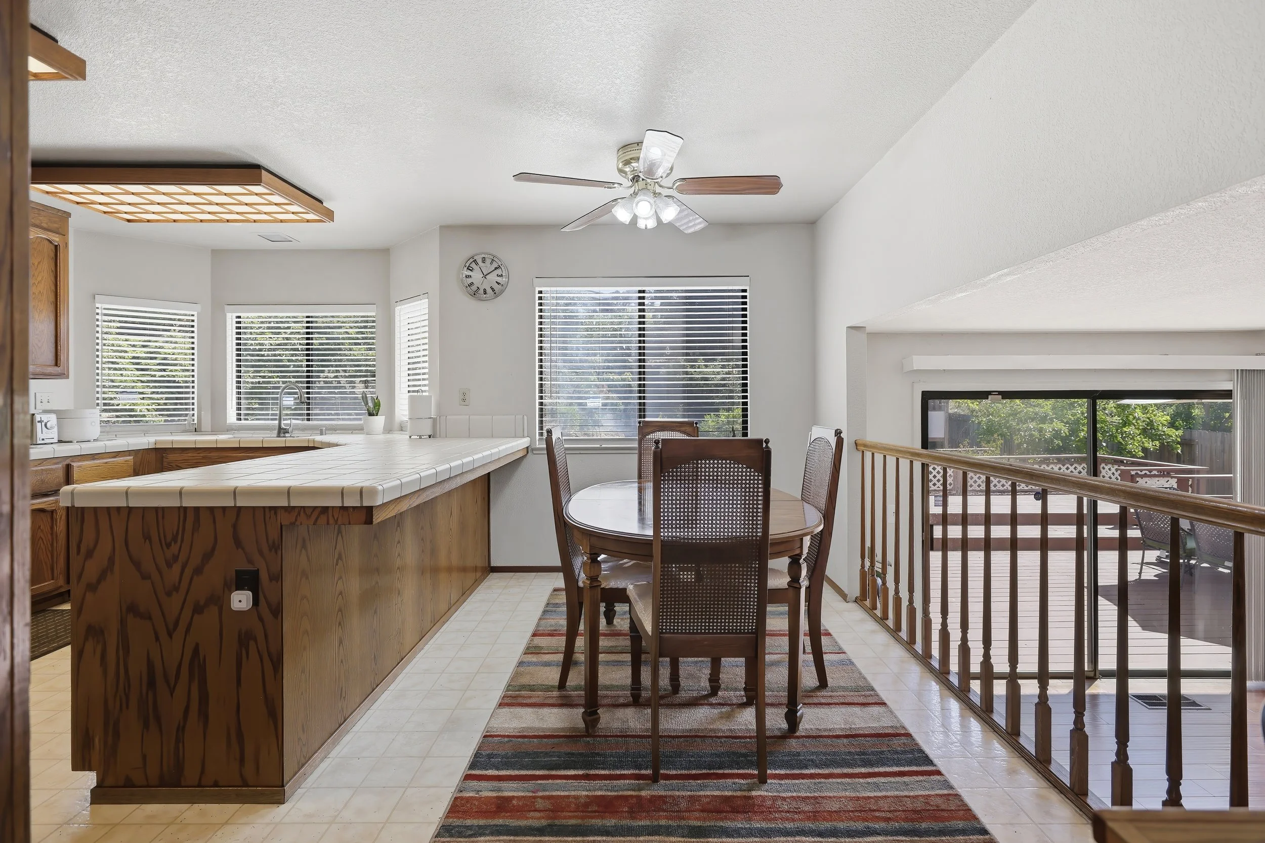 A kitchen and dining area with a white tiled counter, wooden cabinets, large windows with blinds, a round wooden dining table with four chairs, a ceiling fan with lights, and a sliding glass door leading to a deck with trees outside.