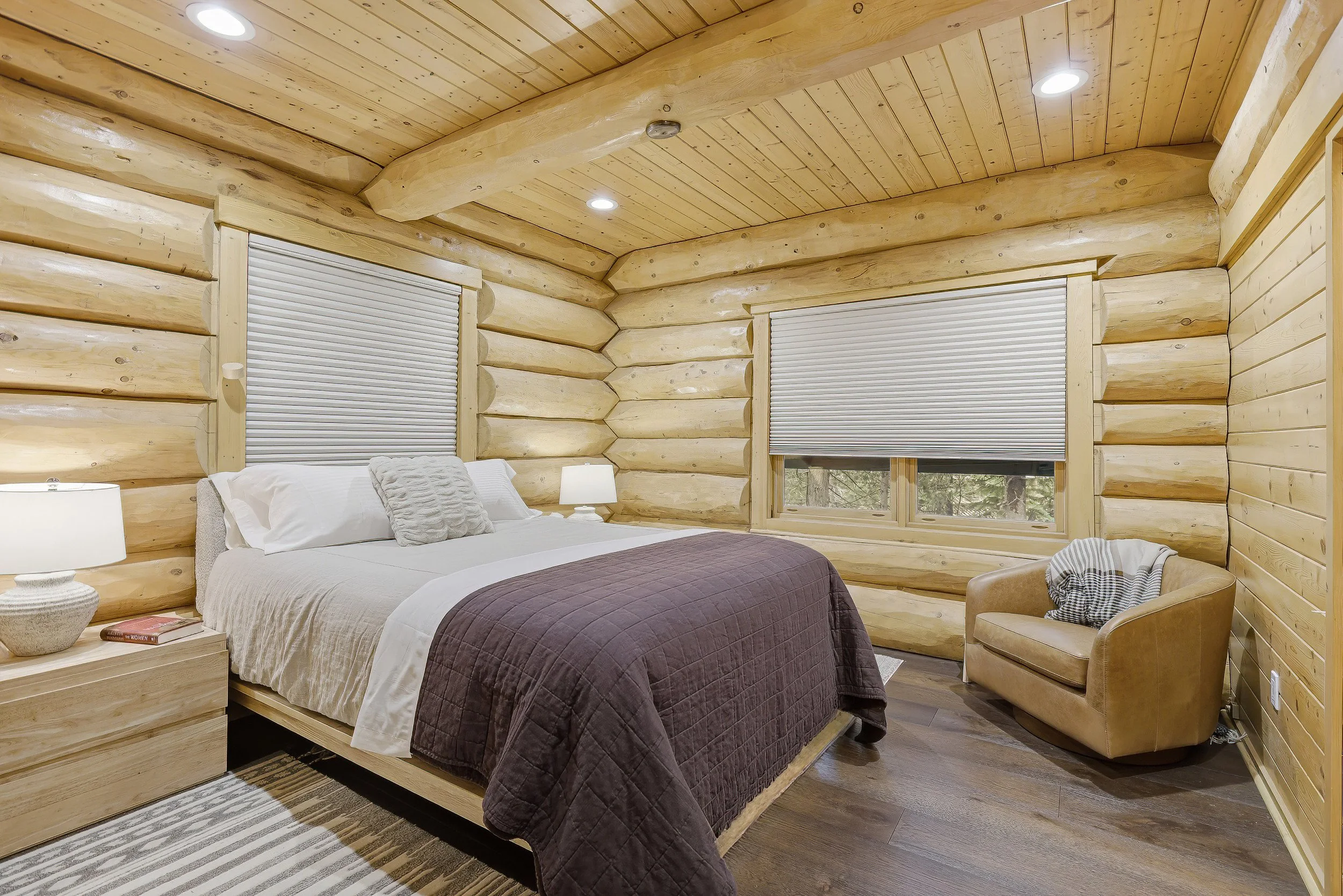 A bedroom with wood log walls and ceiling, two windows with blinds, a bed with pillows and a brown quilt, and side tables with lamps.