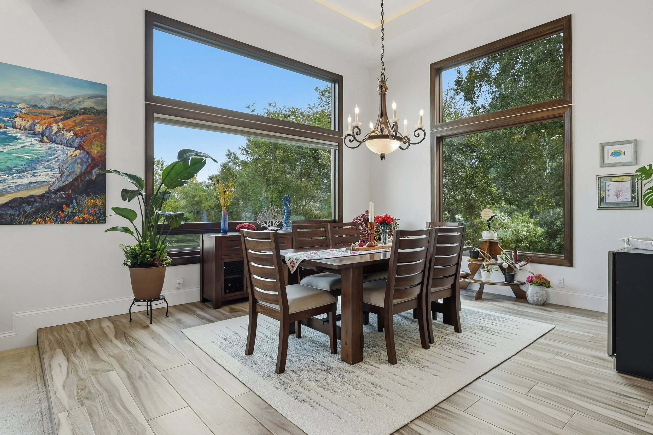 Dining area with wood table, chandelier, and tall windows overlooking greenery.