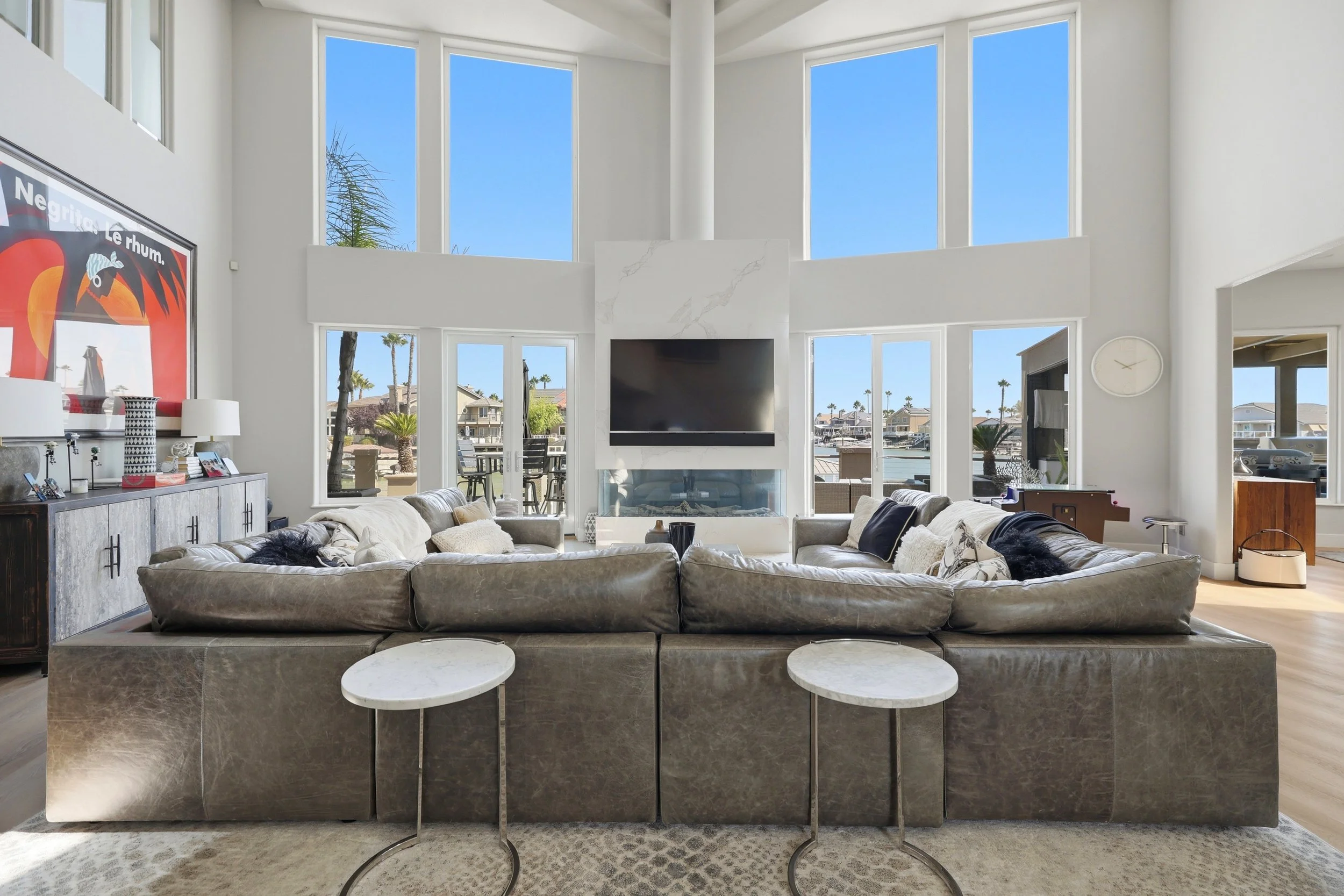 Interior of a modern living room with large windows, a fireplace, a flat-screen TV, a clock on the wall, and a sectional sofa with cushions and blankets.