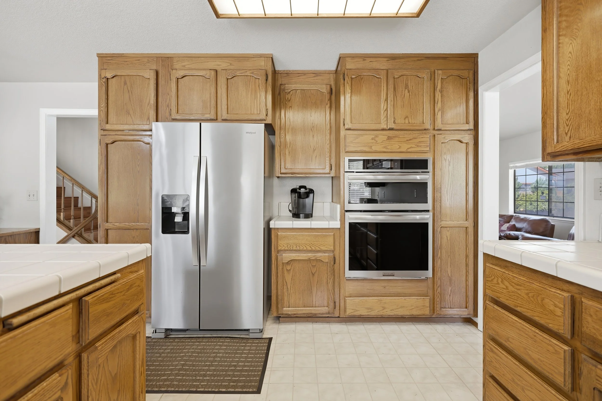 View of a kitchen with wooden cabinets, a stainless steel refrigerator, and a built-in oven. There are countertops, a black coffee maker, and windows on the right side, with a living room visible through an open doorway.
