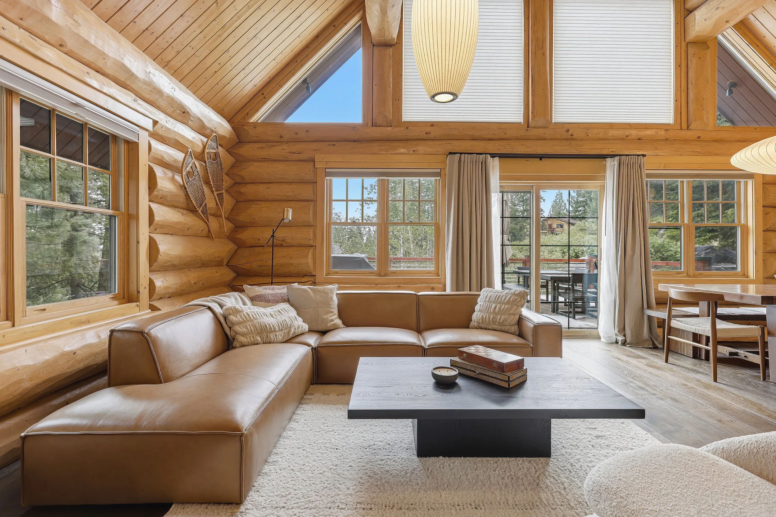 Living room with large windows, wooden interior, beige sectional sofa with pillows, coffee table, and outdoor patio beyond the windows.