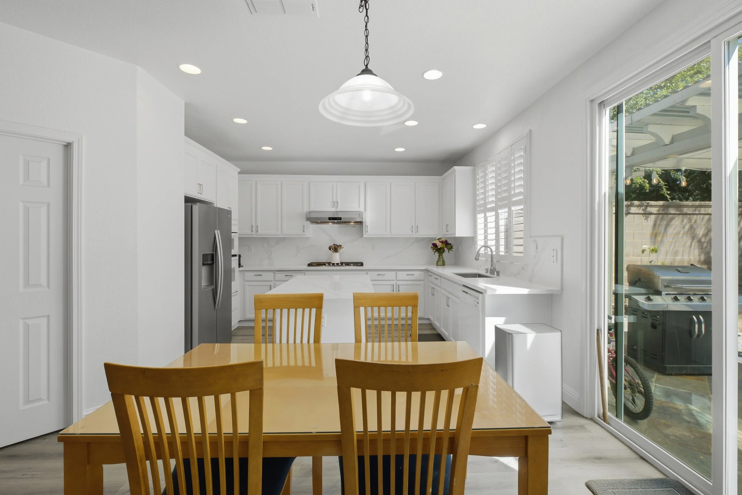 A kitchen with white cabinets, a stainless steel refrigerator, a marble countertop, and a view outside through sliding glass doors showing a patio with a grill and outdoor area. There is a wooden dining table with four matching chairs.