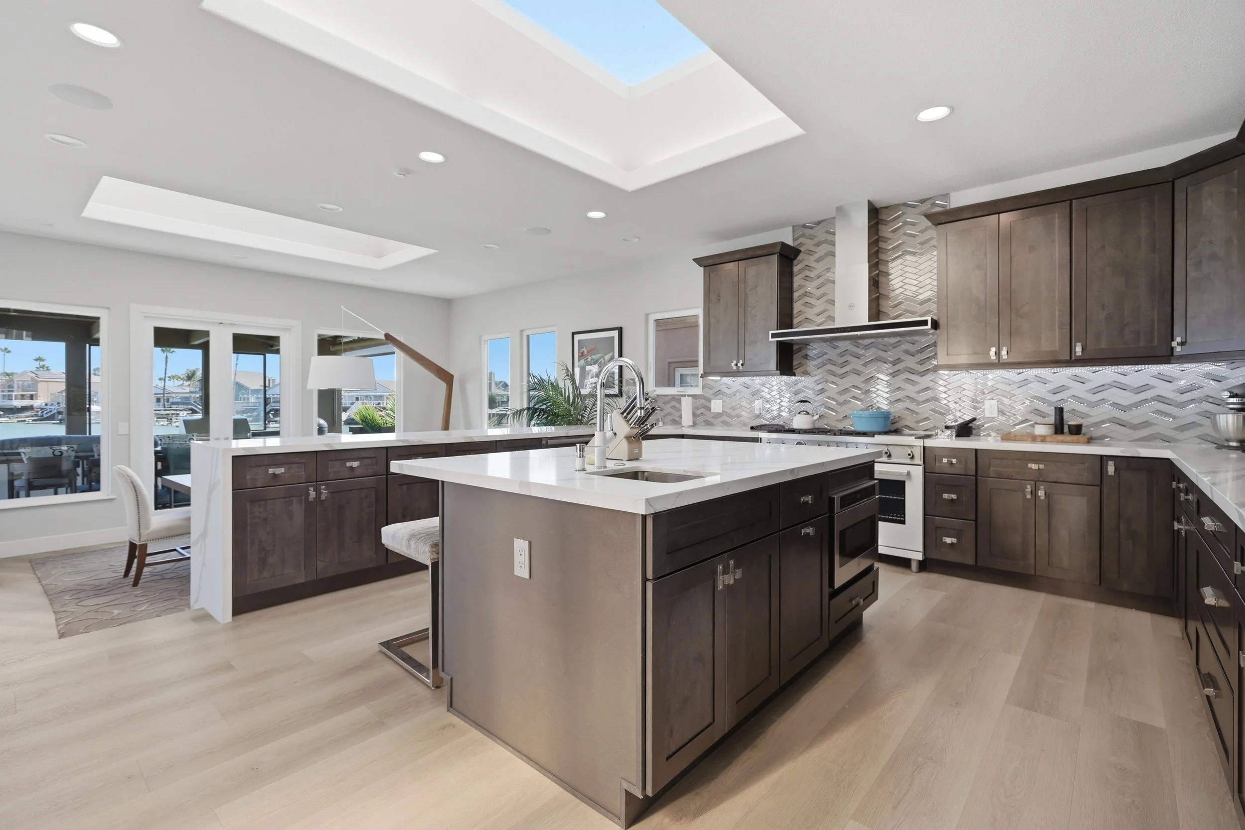 Modern kitchen with dark wood cabinets, white marble countertops, stainless steel appliances, and a backsplash with a chevron pattern. A skylight is visible on the ceiling, and windows overlook a marina with boats.