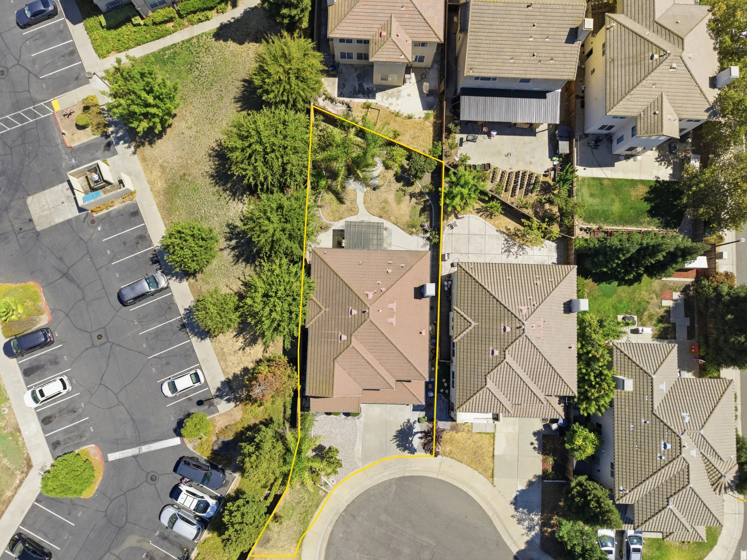 An aerial view of a residential neighborhood with houses, trees, a parking lot, and paved walkways, with a yellow outline marking a specific yard area.