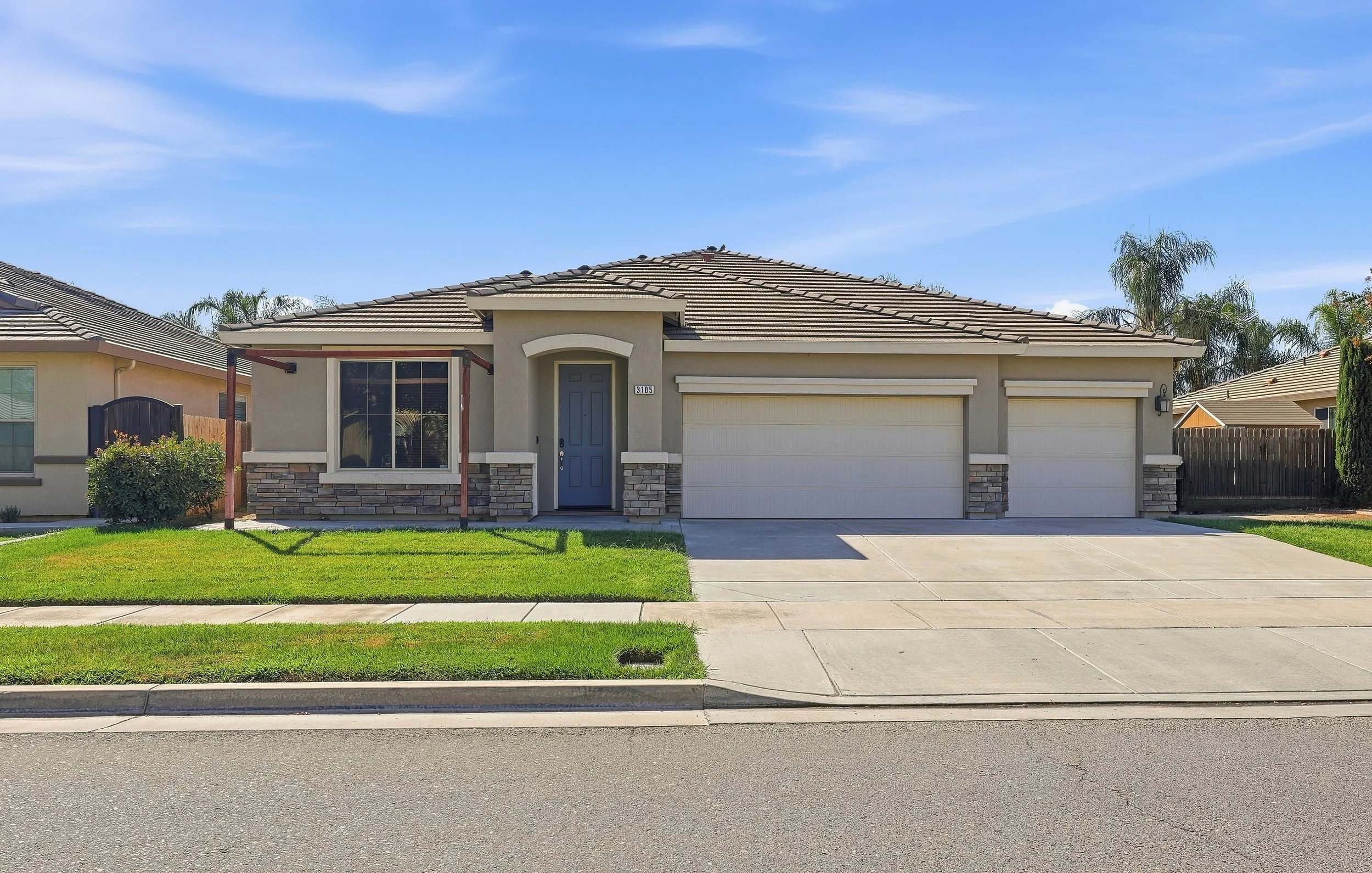 Front view of a modern single-story house with a two-car garage, a blue front door, stone accents, and a small front yard with green grass, on a sunny day with clear blue sky.